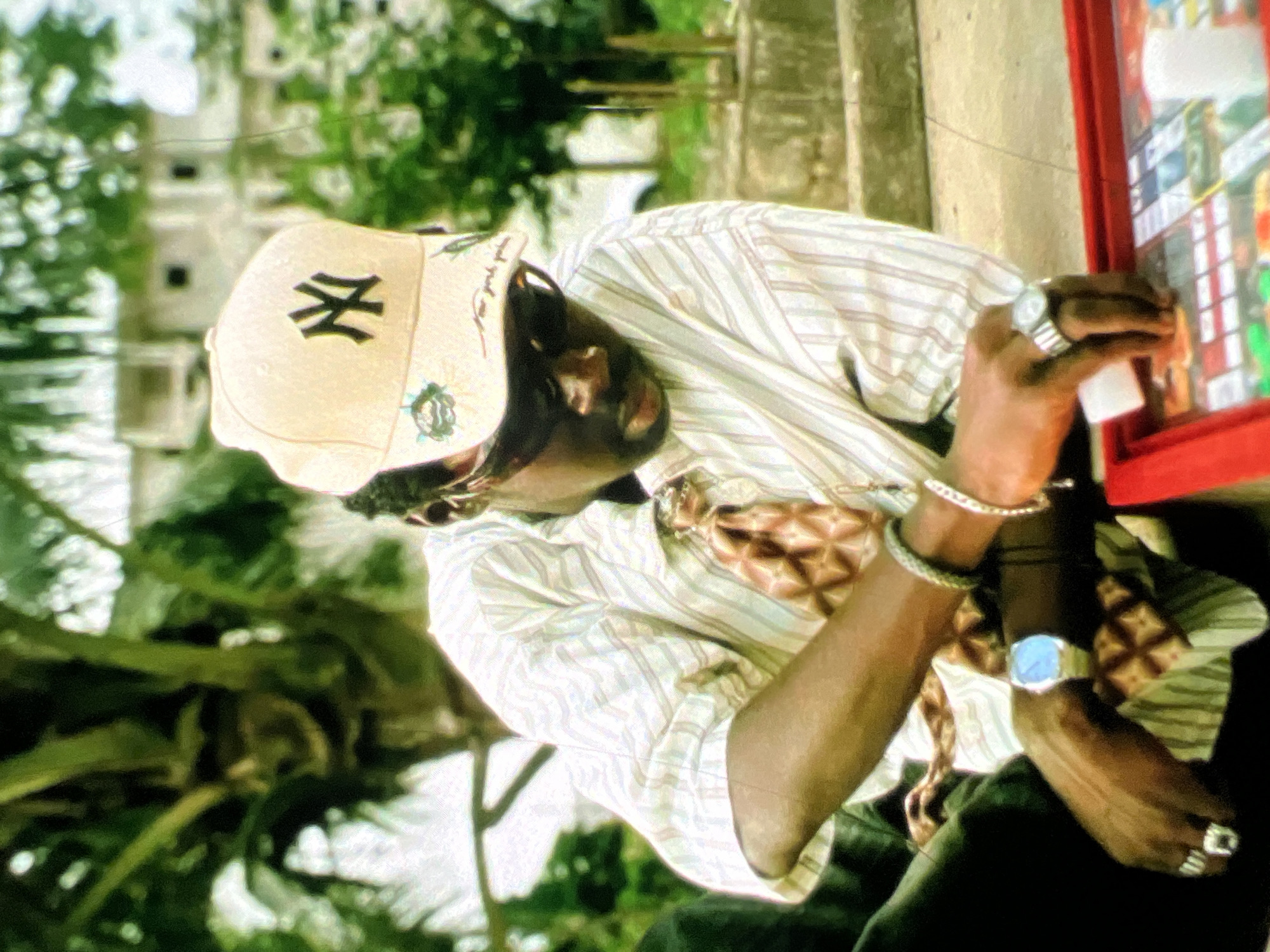A medium shot captures a Black male from the chest up, seated outdoors in an urban environment, possibly Lagos, Nigeria, given the tropical foliage and architecture. He is wearing a tan New York Yankees baseball cap, sunglasses, and a vertically striped collared shirt over a patterned tie. The man is actively engaged with a red board game, his hands positioned over the game's surface. He wears a silver watch on his left wrist and several rings on his fingers. The background features lush green trees and the facade of a building, suggesting a residential or communal outdoor space. The lighting is bright, indicating daytime.