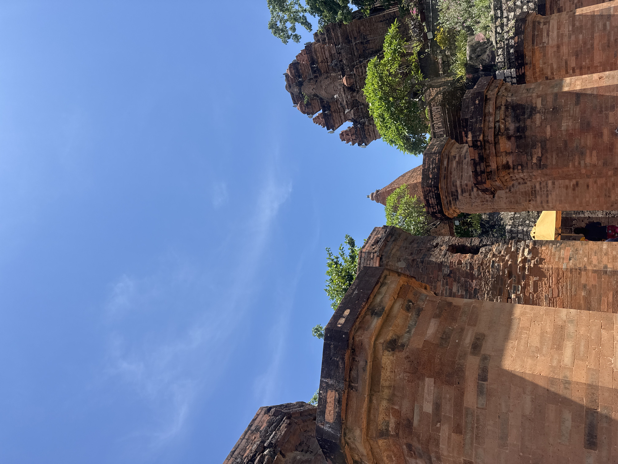 The image shows the Po Nagar Cham Towers in Bac Nha Trang Ward, Vietnam. The sky is a bright blue with a few wispy clouds. The towers are made of red brick and are partially covered in greenery. In the foreground, there are large brick pillars. In the background, a tiered tower is visible, with trees and plants growing around it. A yellow umbrella and a person are visible in the lower right portion of the image. The overall scene depicts an ancient architectural site on a sunny day.