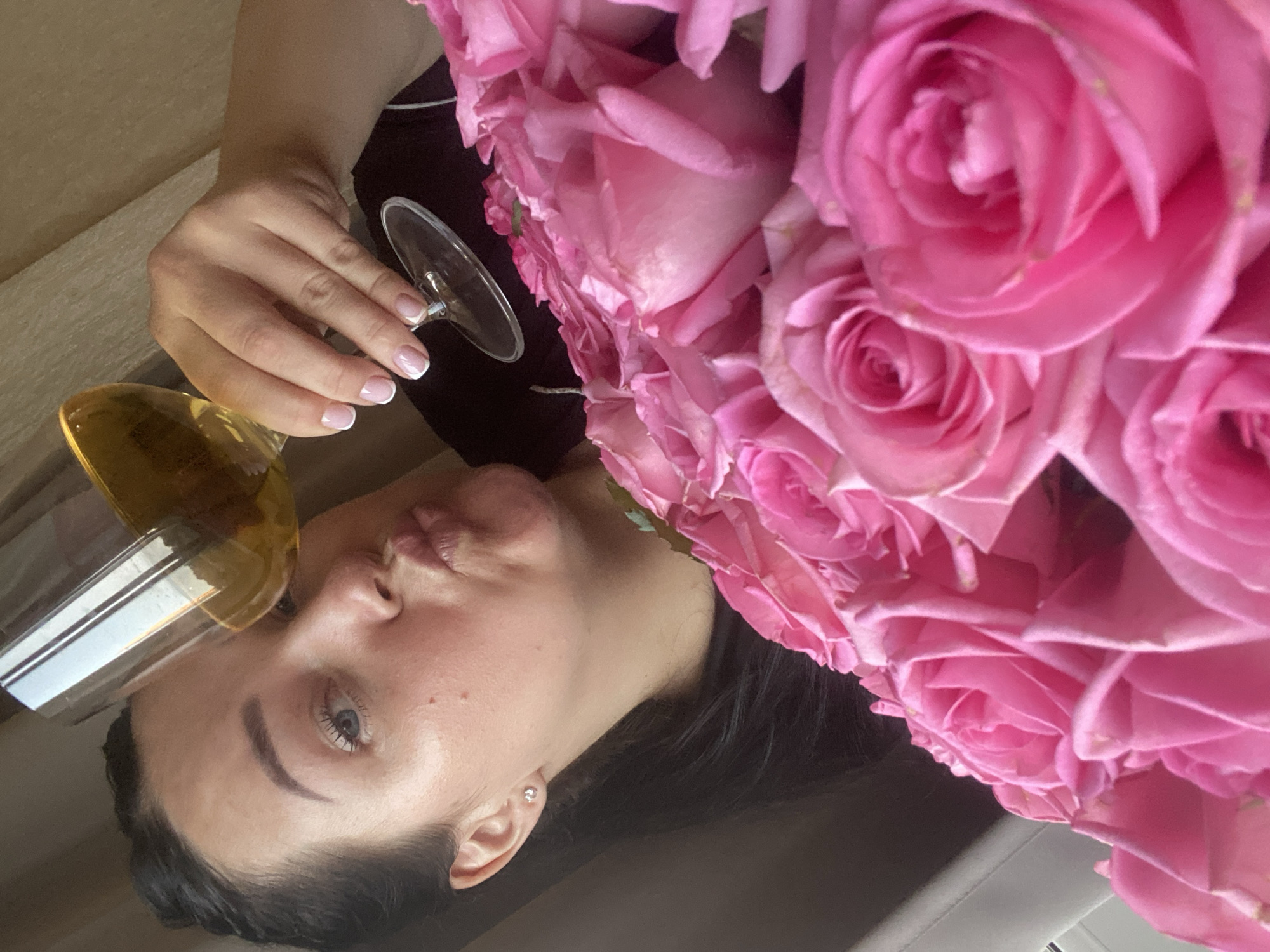 A light-skinned woman with dark hair is oriented at an oblique angle, holding a glass of golden liquid in her left hand, and resting her face near a large bouquet of pink roses. She has white-tipped, manicured nails. The location appears to be indoors, with a neutral-toned background. There is no verifiable information in the image to suggest that the location is in Sochi, Russia.