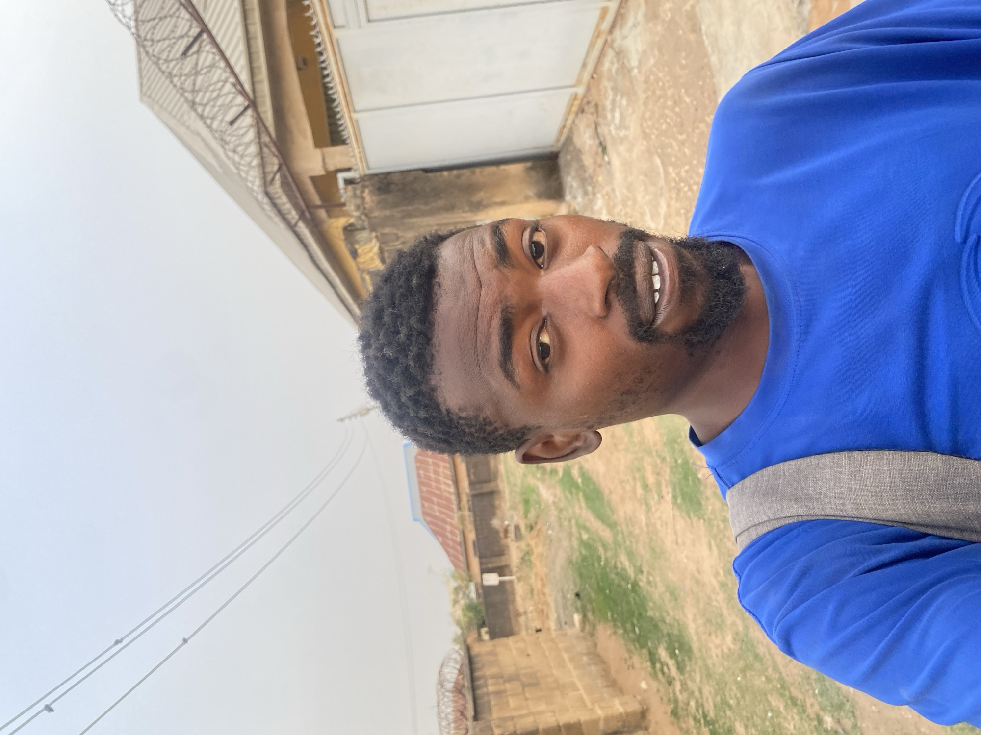 This is a portrait of a young adult Black man taken outdoors in Barakin Akawo, Nigeria. The man, likely in his 20s or early 30s, has dark skin, short curly black hair, and a trimmed beard and mustache. He is wearing a bright blue long-sleeved shirt and a grey strap, likely from a backpack, is visible over his right shoulder. He looks directly at the camera with a slight, friendly smile, showing his teeth.
The setting appears to be a semi-urban or residential area. In the background, there are buildings under what looks like a bright, overcast, or hazy sky, indicating daytime with diffused light. To the man's left, a building with a light-colored upper section and a metal fence, possibly topped with barbed wire, is visible. Further back, another building with a rusty corrugated metal roof can be seen. The ground is dry and dusty in places, interspersed with patches of sparse green grass. Overhead, power lines stretch across the frame. There are no specific events taking place, but rather a posed photograph capturing a moment in a typical outdoor environment. No visible text is present in the image.