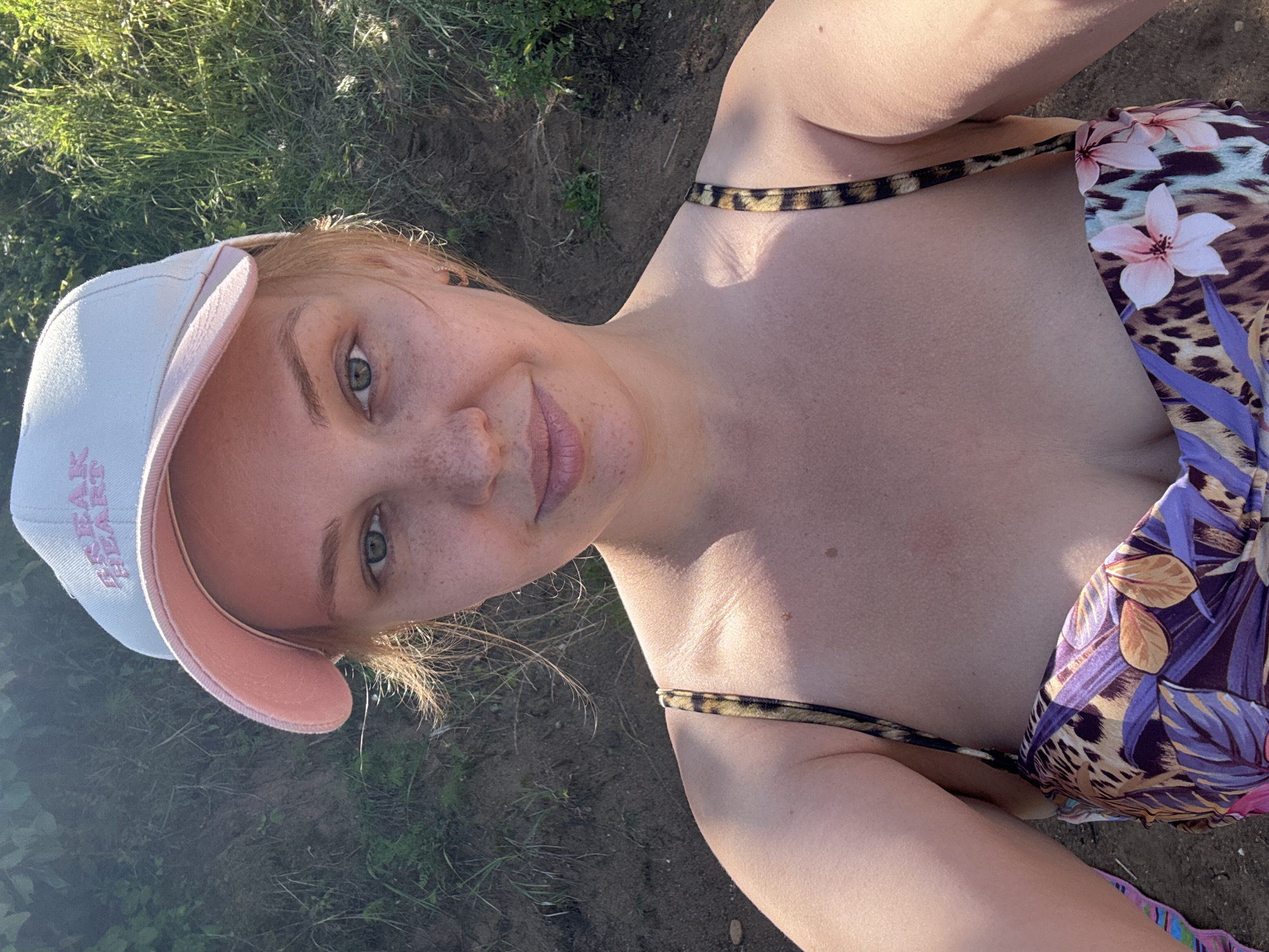 The image shows a selfie of a young Caucasian woman with blonde hair and fair skin, wearing a white baseball cap with pink lettering that reads "BREAK HEART" and a bikini. She has freckles across her nose and cheeks. She is outdoors, with a background of sandy soil and green grass. The sun is shining brightly, creating a strong light on her face and shoulders. She is wearing a bikini top with a leopard print and floral pattern. The overall scene suggests a casual, outdoor setting, possibly a park or beach. The image does not contain any information that would allow for an inference about its specific location in Yaroslavl, Russia, or any other city.