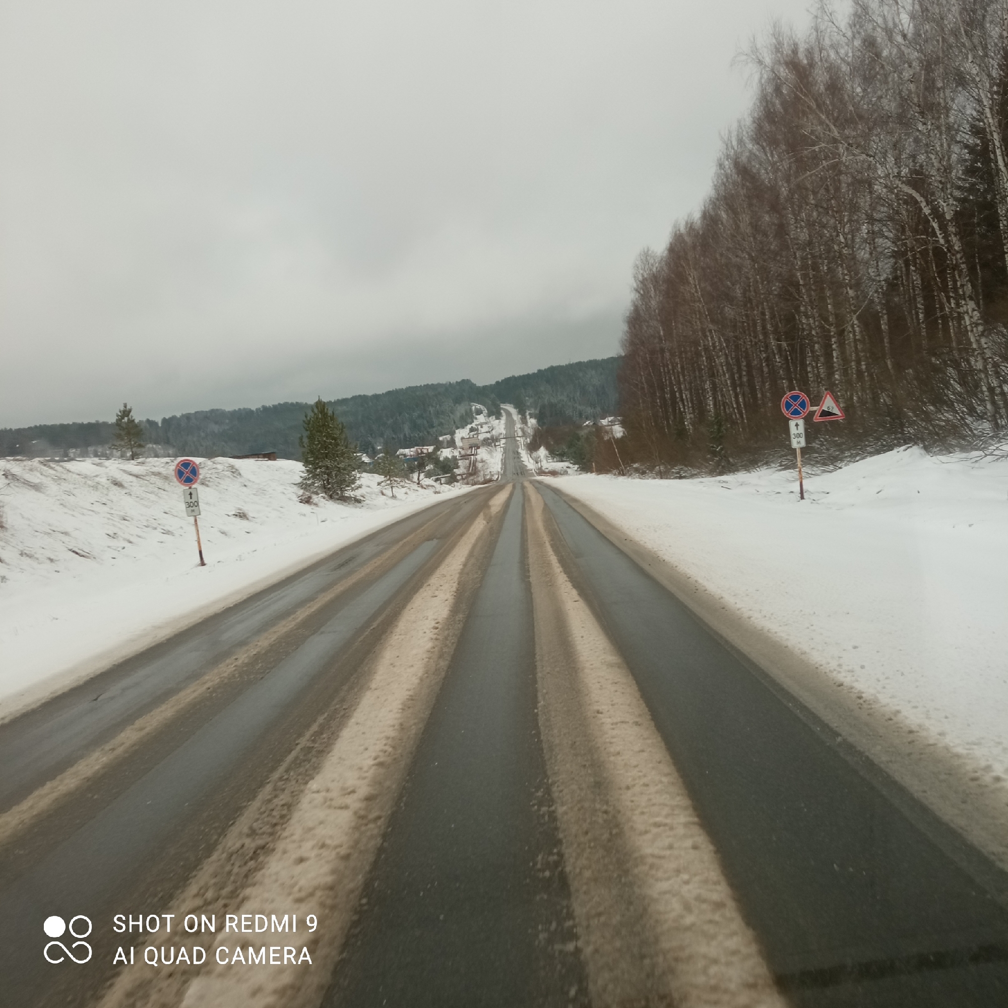 The image captures a snow-covered road in Ольховка, Russia, under an overcast sky. The road, which appears to be asphalt, is marked by parallel lines of melting snow. The shoulders of the road are covered in snow. On either side, there are trees and patches of forest, also covered in snow. In the distance, the road leads towards a hillside with a ski jump. Road signs are visible on the right side of the road. The image includes a watermark indicating it was taken with a Redmi 9 AI Quad Camera.