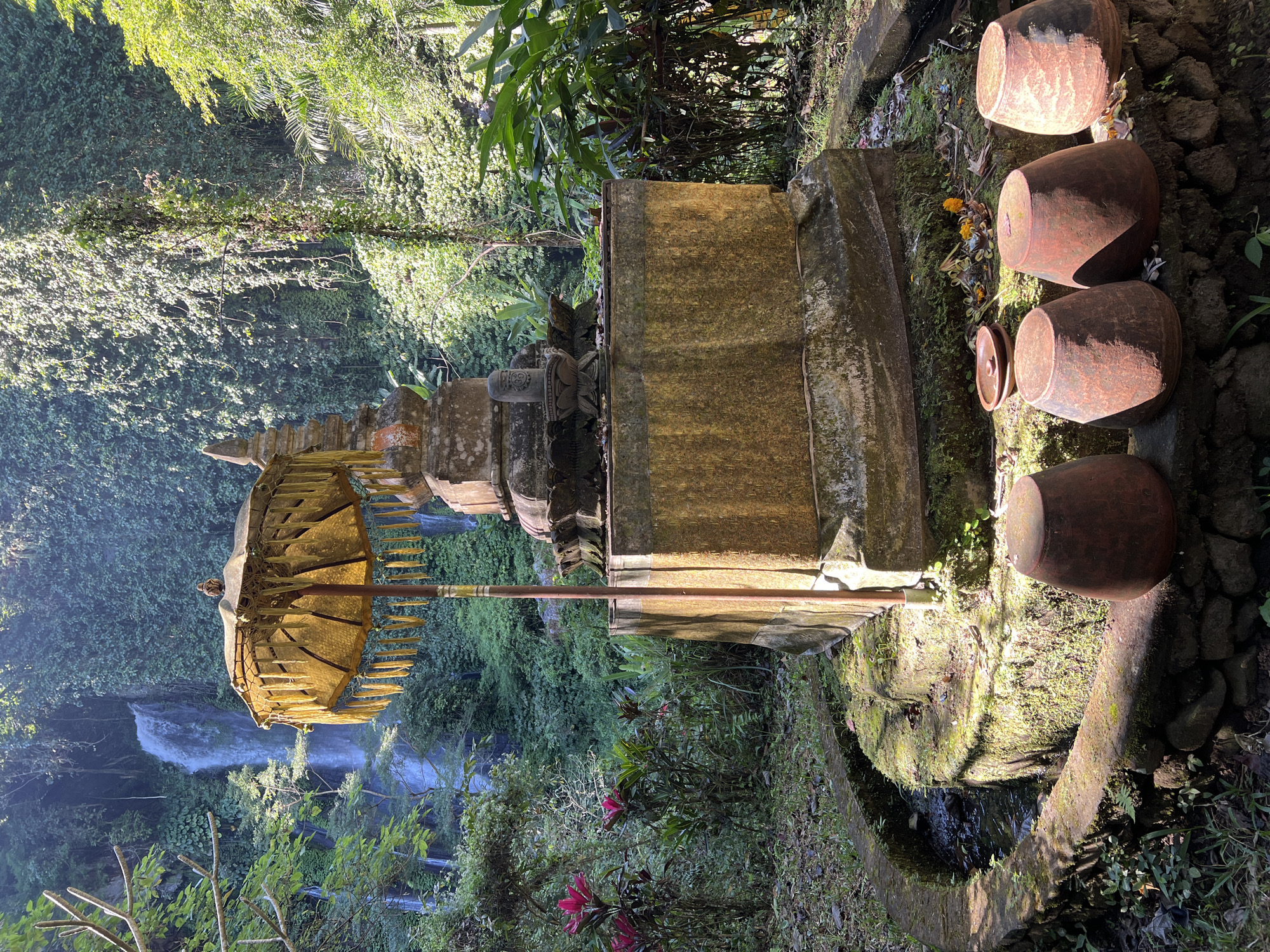 In Busungbiu, Indonesia, a stone altar with an ornate umbrella stands in a jungle setting. A waterfall cascades in the background, surrounded by lush greenery. Several terracotta pots are placed around the base of the altar. The scene appears to be a place of worship or a significant cultural site.