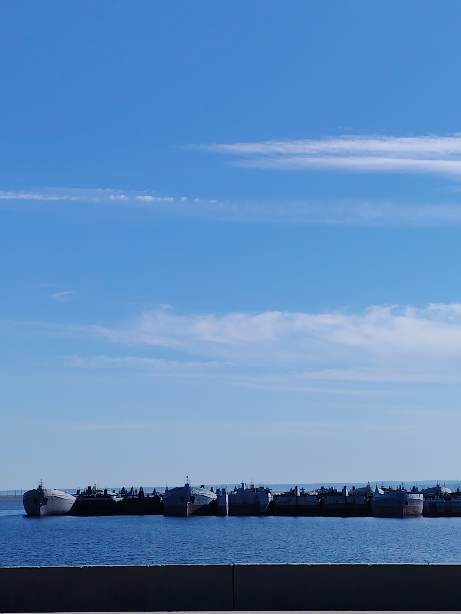The image shows a cluster of inactive ships anchored in the Votkinsk Reservoir, a body of water near the city of Chaikovsky, Russia. The sky is clear and blue with scattered thin clouds. The ships are of varying sizes and appear to be in a state of disuse. In the foreground, there is a concrete structure.