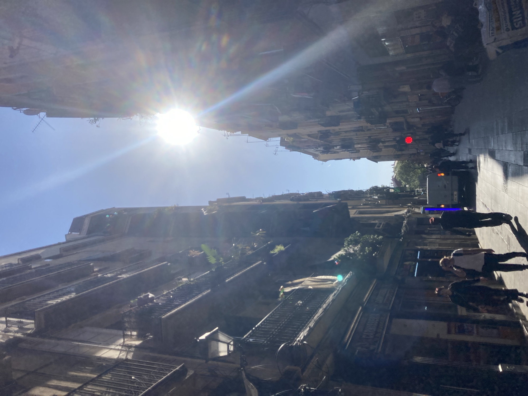 A vibrant street scene captured from a low angle, looking along a narrow urban street lined with multi-story buildings. The bright sun, positioned in the upper left, creates significant lens flare and strong glare, casting deep shadows across much of the scene. The sky is clear blue.
On both sides of the street, tall buildings feature numerous balconies, some adorned with plants. The building on the left has distinctive external metal staircases or fire escapes. On the right, pedestrians are visible on the sidewalk, their forms often silhouetted against the bright light or obscured by shadows. One person in a white shirt and dark pants is clearly seen walking forward on the right.
Notable details include a prominent red traffic or pedestrian signal positioned high on the right side of the street. The pavement appears to be tiled or cobbled. Shopfronts line the ground floor of the buildings on the right.
Visible text includes partial signs such as "SINKS" and "R-5" towards the top right. More clearly, a shop sign on the right reads "CARNICERIA" (butchery) and "CARRER DE S. LLORENÇ" (Saint Lawrence Street), indicating a location in a Catalan-speaking region. The intense sunlight and long shadows suggest it is either early morning or late afternoon.