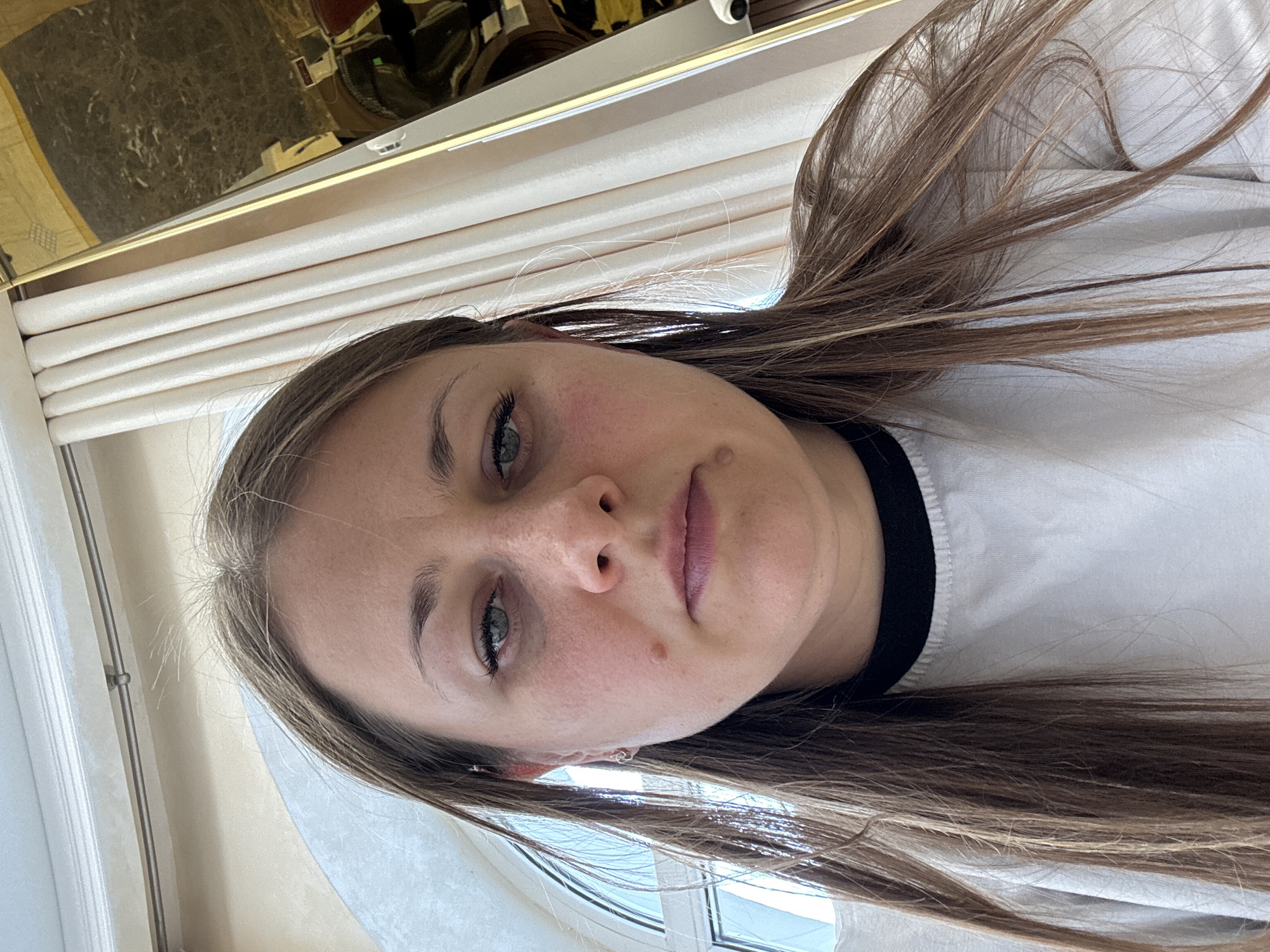 A close-up, selfie-style photograph captures a woman's face from a low angle. She has light brown hair, which is parted and falls around her face and shoulders. Her eyes are open, and her gaze is directed towards the camera. She is wearing a white long-sleeved shirt with a black collar. The background suggests an indoor setting with light-colored curtains and a window with an arched top on the left side. A portion of a gold-colored ornate mirror is visible on the right, reflecting a dark, patterned surface and some objects. The overall lighting is bright, with some shadows. The woman has a mole on her left cheek and appears to have light makeup on her eyes and lips. The scene does not contain any identifiable landmarks or features that would place it specifically in Yevpatoriya, Ukraine.