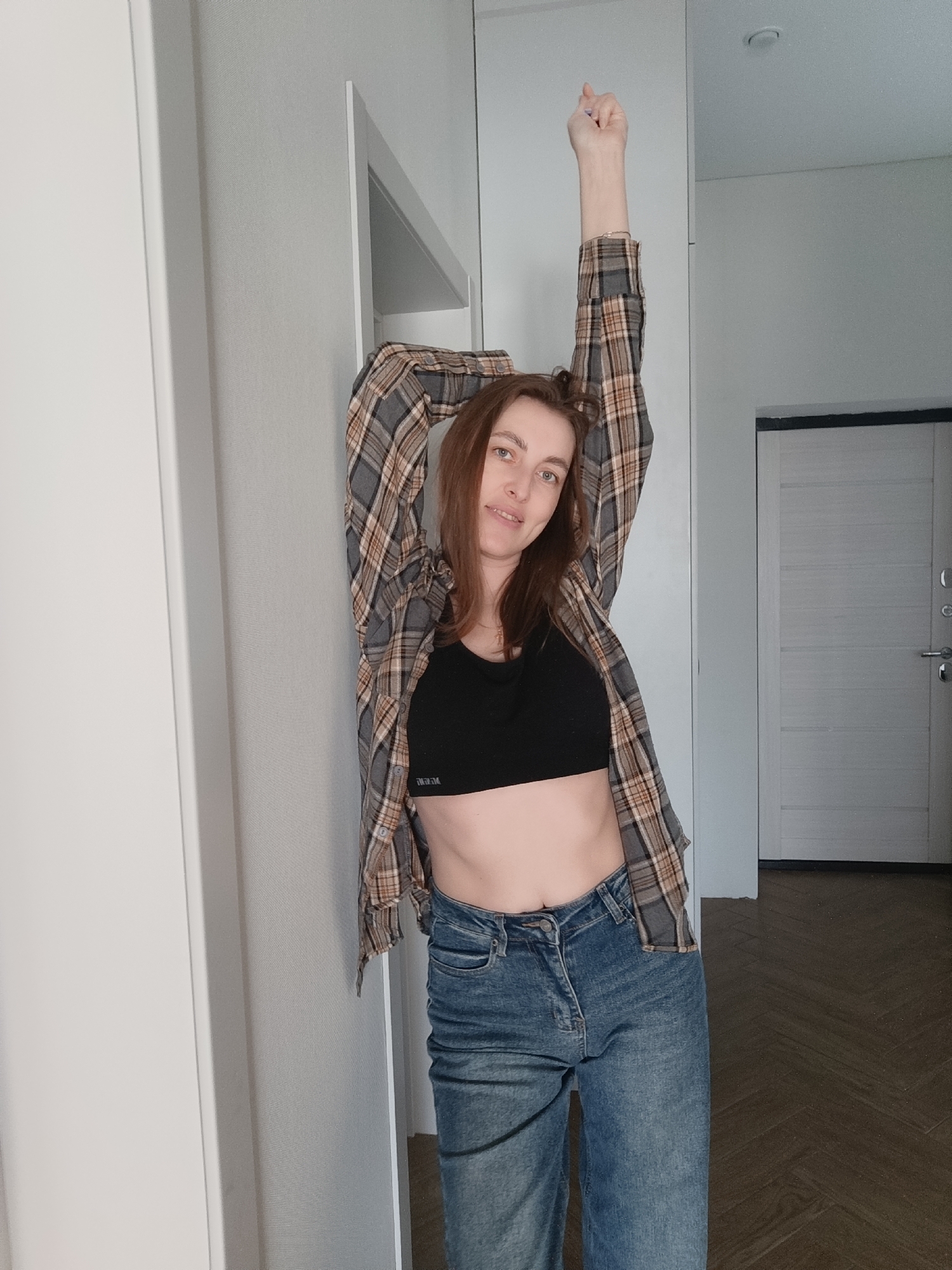 A young woman stands in what appears to be an indoor hallway or entryway. She is wearing a black sports bra, blue jeans, and a plaid shirt that is open. One of her arms is raised above her head, while the other is slightly bent and resting against a wall. The walls are white. There is a white door behind her to the right and a doorway to the left. The floor is a dark wood grain. It is unknown if the image was taken in the city of Chaikovsky, Russia.