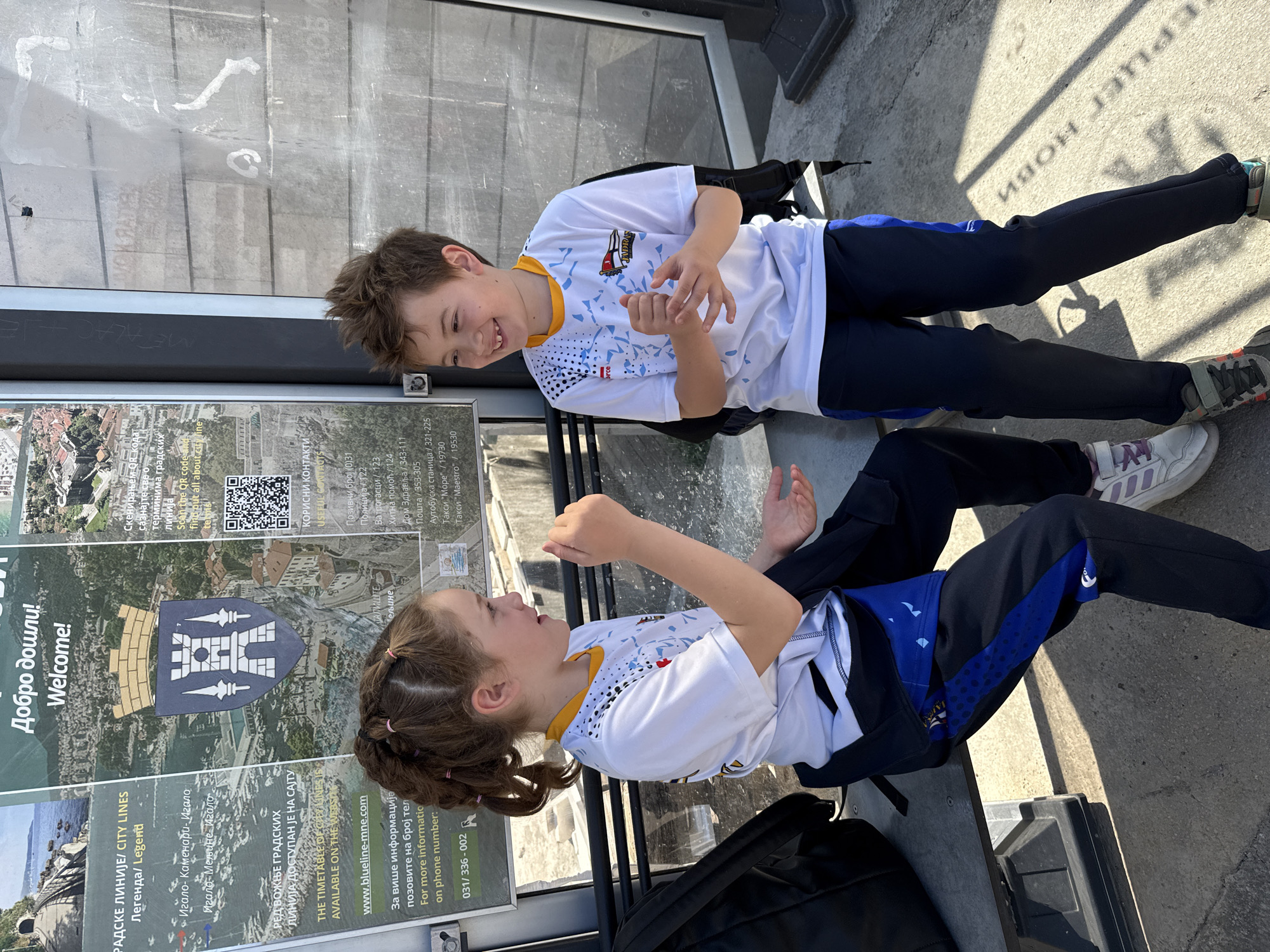 Two children, a boy and a girl, are seated on a bench in Meljine, Montenegro. They are both wearing white shirts with blue and yellow accents, along with dark-colored pants. They are facing each other and appear to be engaged in a playful interaction, possibly clapping or making hand gestures. The girl has her hair pulled back in a ponytail. Behind them is a bus stop display featuring information about the local city lines. The bus stop signage includes text in Cyrillic and English, stating "Welcome!" and details about bus routes, contact information, and a QR code. The signage is related to the city lines in Herceg Novi, Montenegro, with information about the local bus lines.