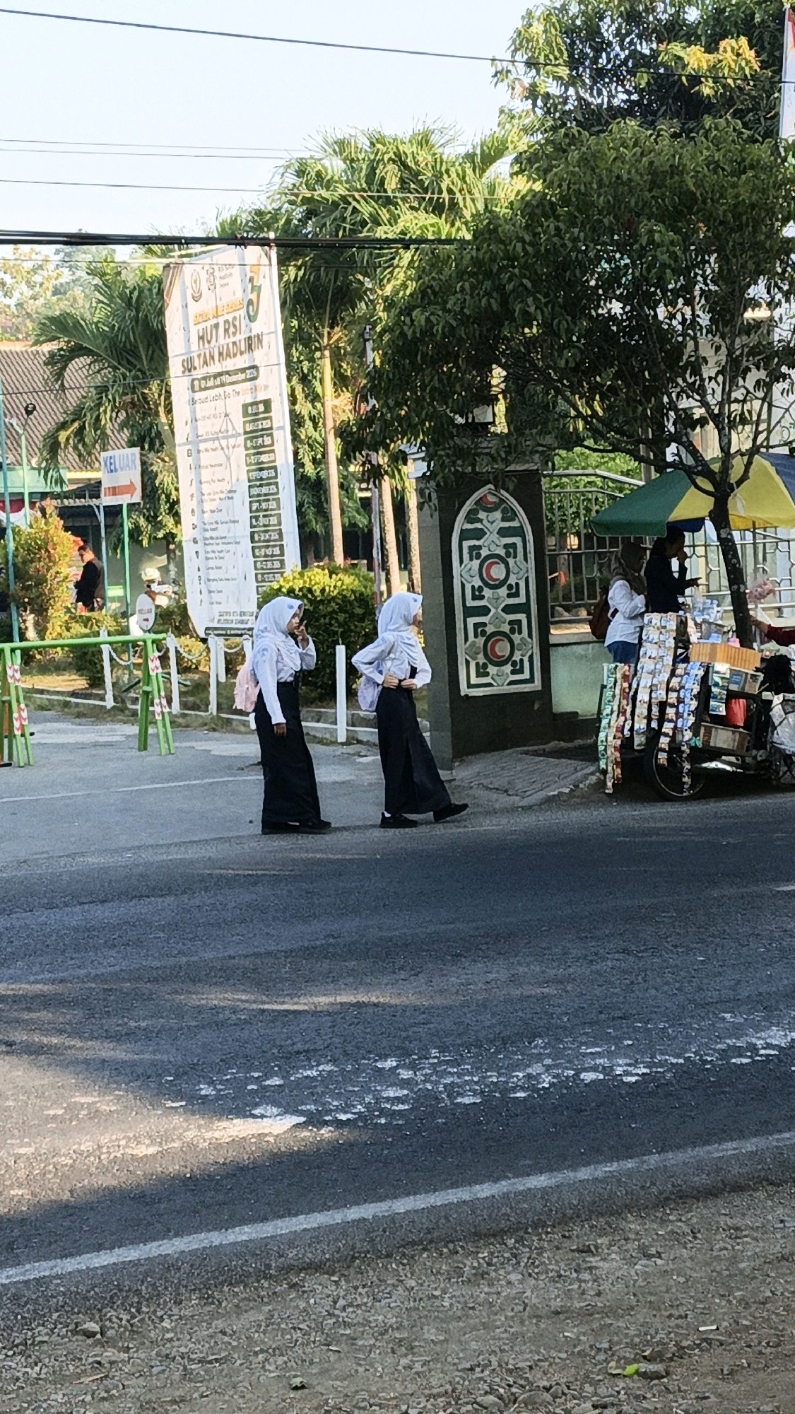Two young women, both wearing white headscarves and long black skirts, are walking across a paved road. The woman on the left is facing towards the right side of the image and appears to be looking down or at something on the ground. The woman on the right is facing towards the viewer and has her right hand raised, seemingly making a peace sign or gesture. They are walking in front of a green gated entrance that has a decorative white and green pattern. To the right of the entrance, a street vendor is operating from a cart under a yellow and green umbrella, selling various items. In the background, there is a tall banner with text in Indonesian, and palm trees and other foliage. The overall scene appears to be a public street in Kuwasen, Indonesia, possibly near an institution or event given the banner.