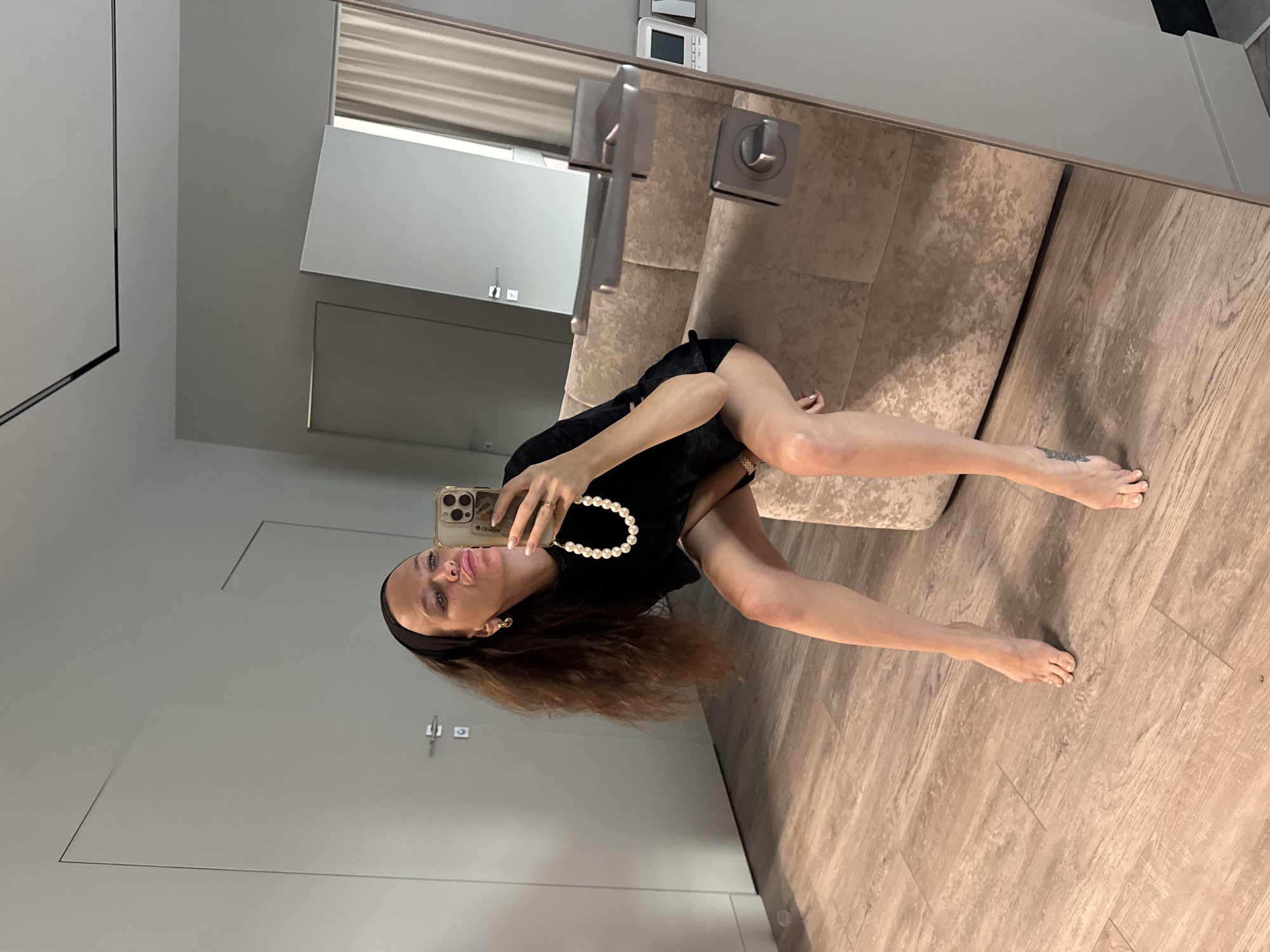 A woman is taking a selfie in a modern, sparsely decorated room. She is sitting on a beige upholstered chair, with her legs crossed and her feet bare. The woman is wearing a black dress, a black headband, and a pearl necklace. She is holding a smartphone in her right hand and making a kissing face. The room has light-colored walls, wooden flooring, and a large mirror that reflects the woman and part of the room. There are doors and a curtain visible in the reflection. A light switch and intercom panel are mounted on the wall near the mirror. The overall impression is one of contemporary urban living.