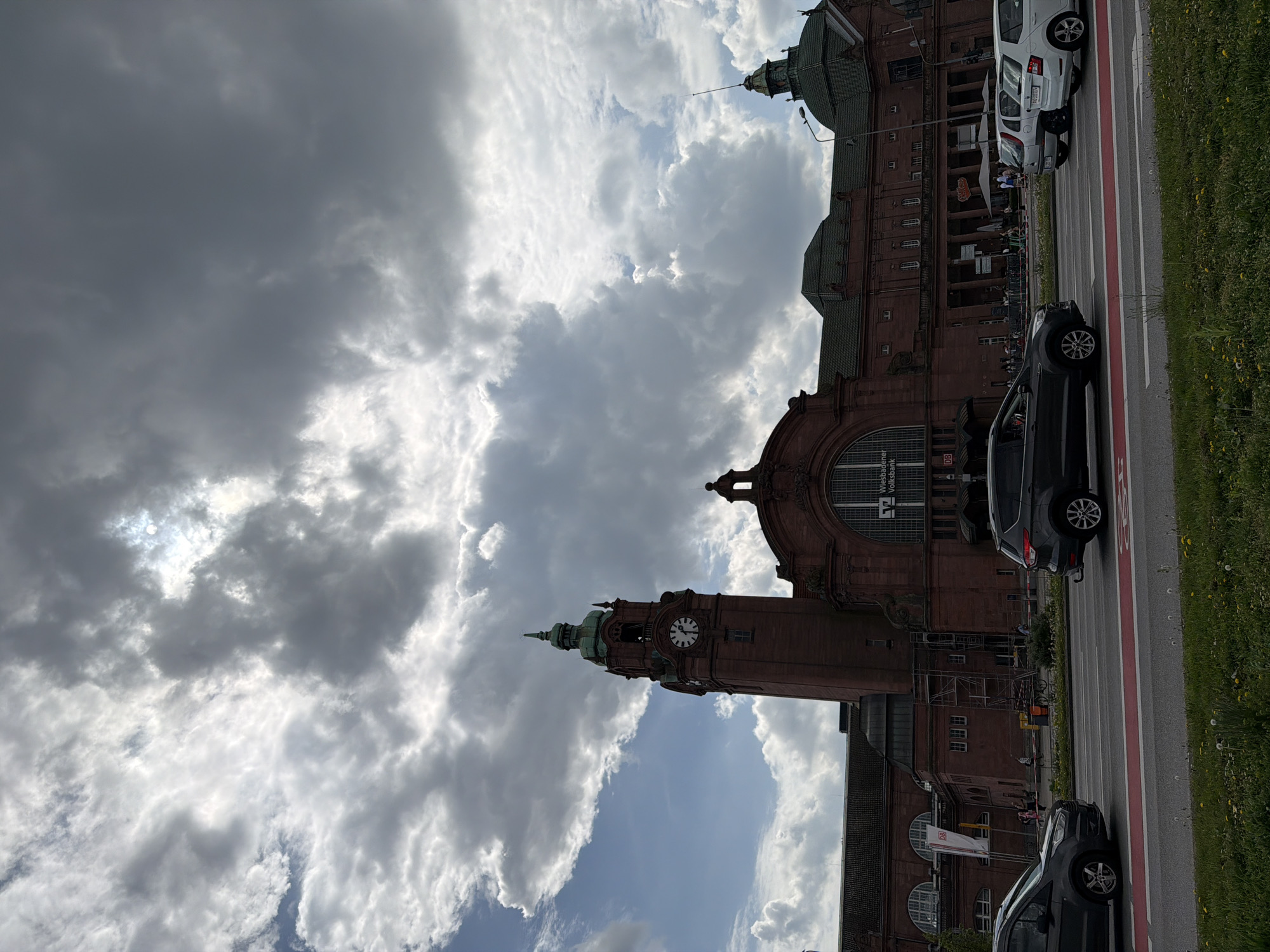 The photograph captures the Wiesbaden Hauptbahnhof, the main railway station in Wiesbaden, Germany. The building is constructed of red brick with a large arched window and a clock tower. The sky is filled with dramatic, partly cloudy conditions. In the foreground, cars are parked on the side of the road next to a strip of grass.