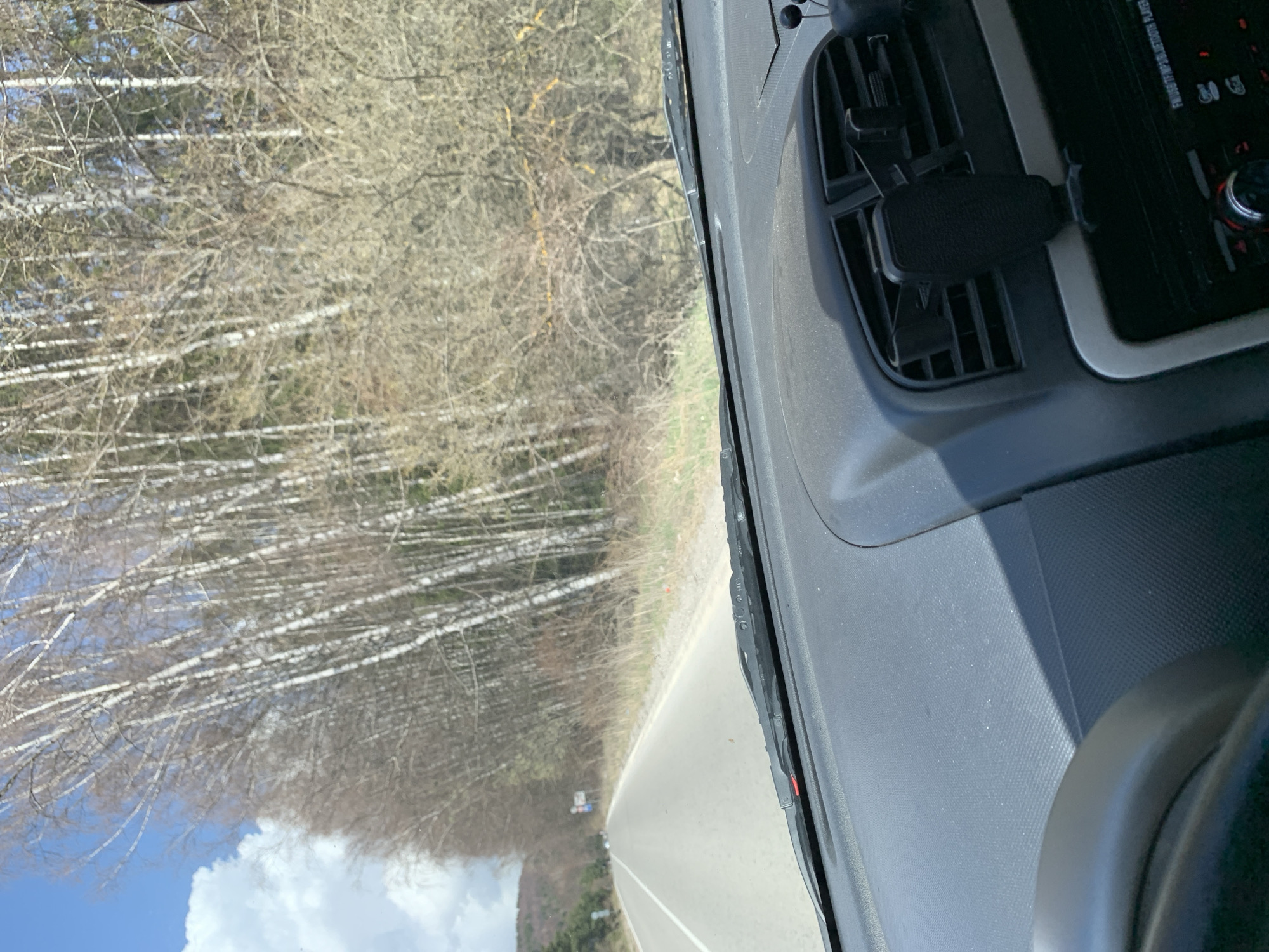 The image, taken from inside a vehicle, shows a road stretching into the distance through a forested area, likely captured during late winter or early spring. The perspective suggests the photographer is a passenger or driver looking out the front windshield.
In the foreground, the dark grey dashboard of the car is visible on the right side, partially obscuring the view. A black phone holder is clipped onto an air vent on the dashboard. Above it, a car stereo is visible, displaying text like "FM/USB/TF/MP3/BLUETOOTH PLAYER" and "BAND/AMS". Fine dust particles are scattered across the dashboard's textured surface.
Beyond the windshield, a paved road with a solid white line on its right edge leads forward. On the left side of the road, a dense stand of trees with characteristic white bark, possibly birches, dominates the landscape. These trees are mostly bare, their branches forming intricate patterns against the bright blue sky, which features scattered white clouds. The ground beneath the trees is covered in dry vegetation. On the right side of the road, sparser bare trees and undergrowth are visible. In the far distance, faint outlines of hills or mountains can be seen under the sky. The bright sunlight indicates it is daytime with clear weather. A small, indistinct road sign is visible in the distance on the left side of the road. The scene depicts a drive through a rural or mountainous region, consistent with the provided context of Gintsi, Bulgaria.