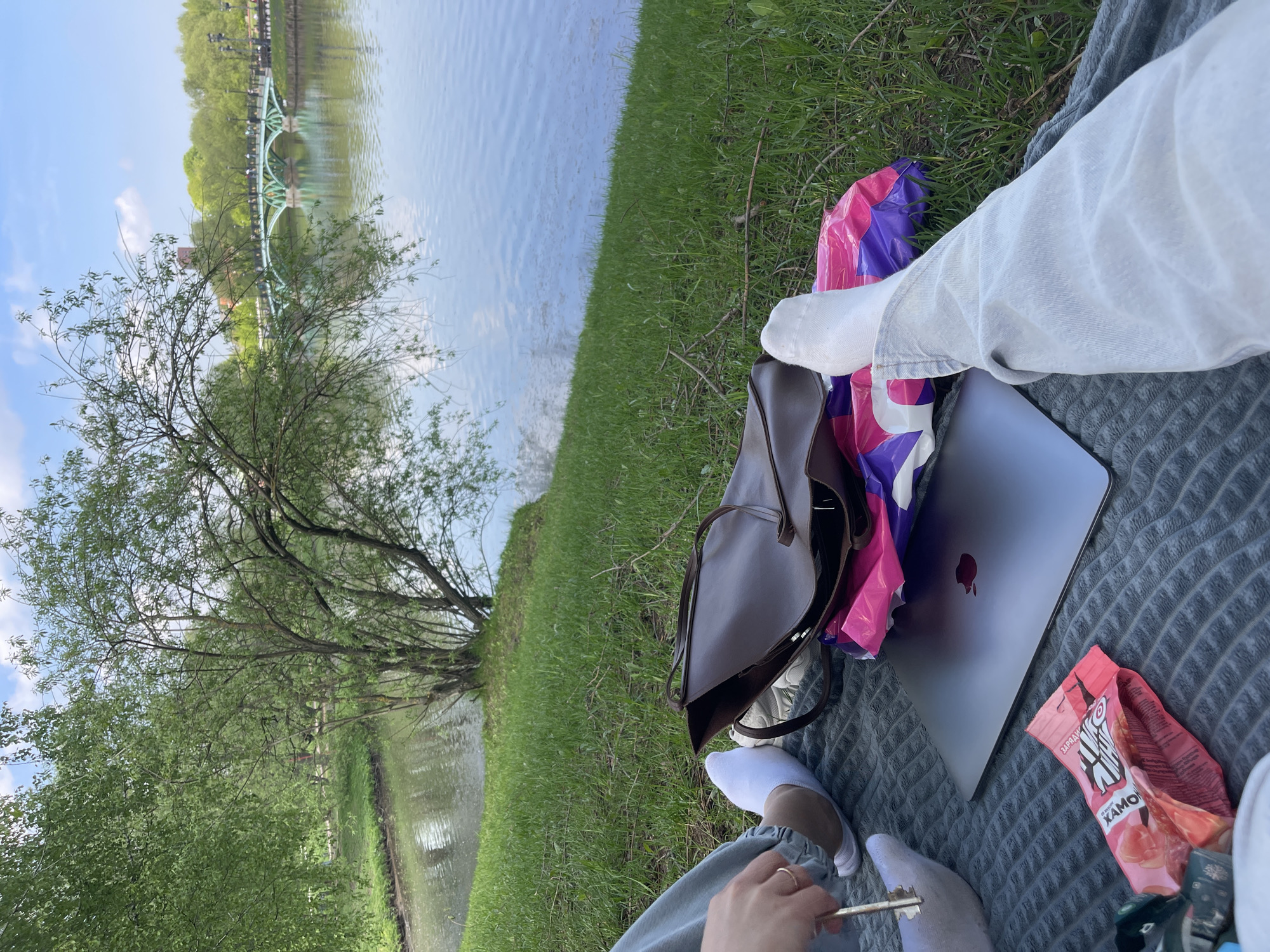 The image depicts a scene outdoors near a body of water, likely a pond or lake, with a view of a bridge in the background, suggesting the location is a park or recreational area. A person is seated on a gray textured blanket laid on the grass near the water's edge. The person is wearing white socks and light-colored pants. A bag, a laptop (likely a Macbook based on the Apple logo visible on the case), and a bag of snacks are also placed on the blanket. The background includes trees and greenery, along with the body of water. The bridge in the distance suggests a location in Moscow, Russia, specifically likely the Tsaritsyno Park due to the architectural style of the bridge visible in the background. The overall scene suggests a relaxing or working environment outdoors.