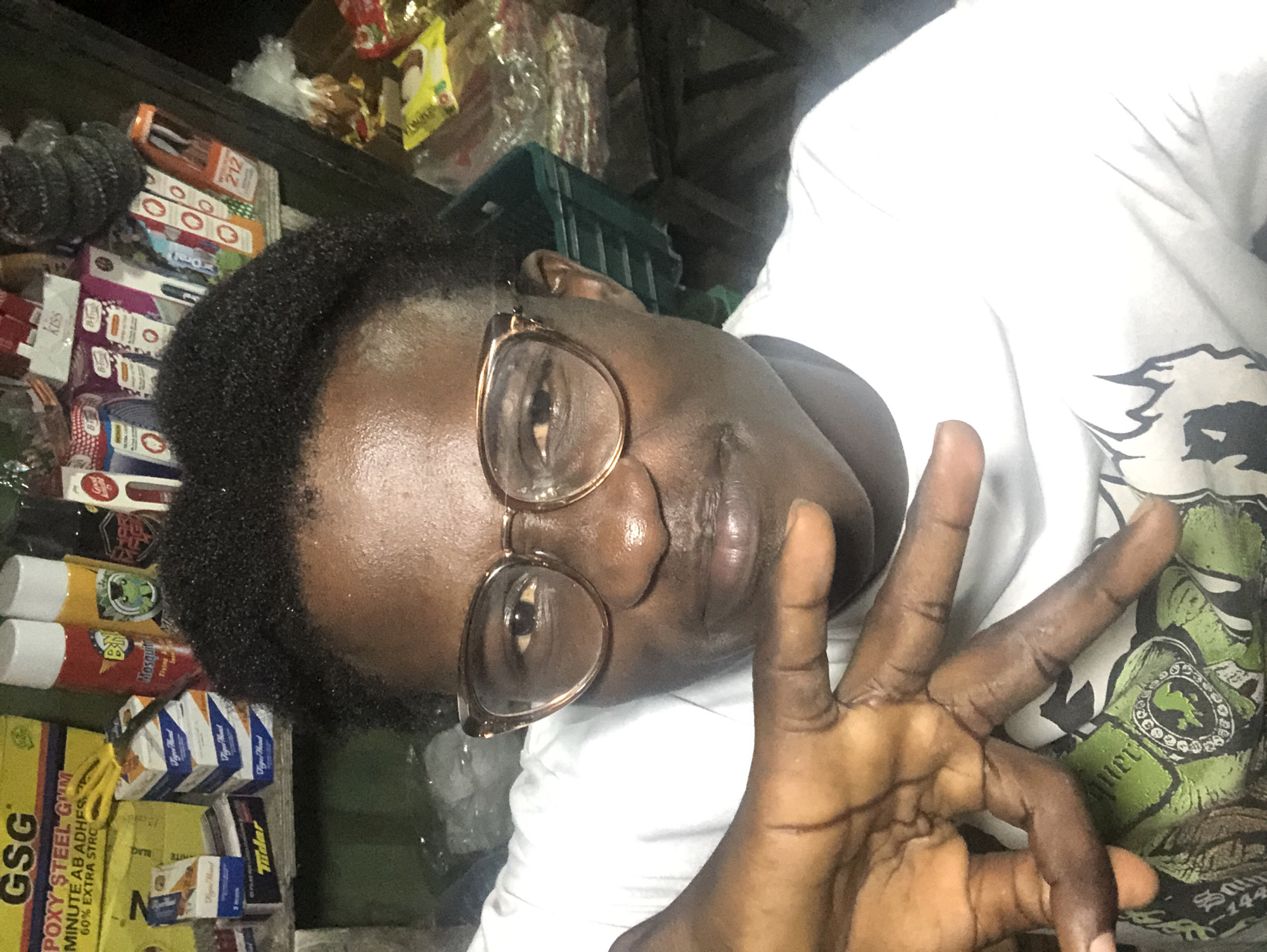 This is a selfie taken indoors, likely in a market stall in Ado, Nigeria. The subject, a young Black male wearing glasses and a white t-shirt, is the primary focus. He is looking at the camera and has his left hand raised, with fingers extended in a gesture. Behind him, shelves are filled with various products, including aerosol cans (one red and one yellow), boxes of what appear to be cigarettes or gum, and other packaged goods. The lighting is dim, casting shadows and highlighting the textures of the products and the subject's skin. The background suggests a commercial setting, possibly a retail stall or small shop, with a green crate visible on the right.