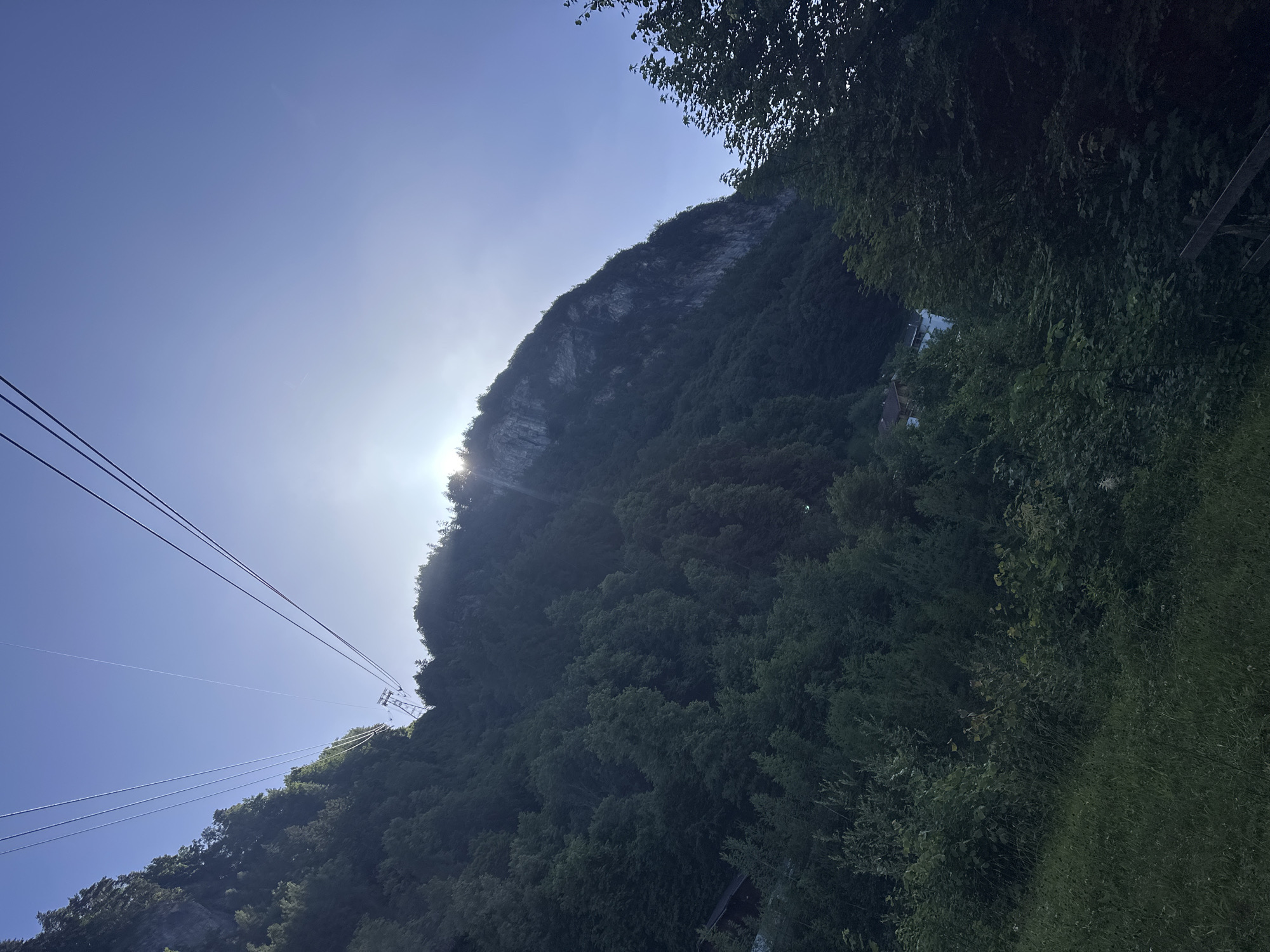 A wide-angle, low-angle shot captures a steep, tree-covered mountainside under a clear blue sky. The sun shines brightly, partially obscured by the mountain, creating a lens flare. Four parallel cable car lines ascend diagonally from the lower left to the upper right of the frame, leading to an unseen station. One cable car structure is visible mid-way up the cables. The mountain is densely forested, with some rocky outcrops visible near the summit on the right side. In the lower left quadrant, a small wooden building is nestled amongst the trees. The foreground consists of a grassy slope. The scene is set in Hüsen, Switzerland.