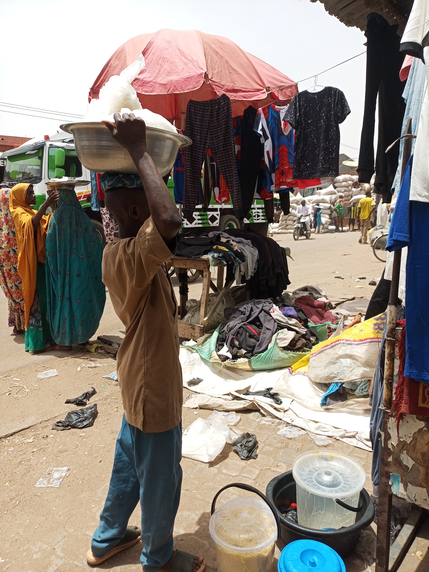 A medium shot reveals a bustling street scene in Arawa, Nigeria, characterized by commerce and daily life. A young boy stands prominently in the foreground, balancing a large, silver metal bowl filled with white, cloud-like material on his head, his hand steadying the load. He wears a brown, short-sleeved shirt and blue pants, with simple sandals on his feet.
To his left, two women stand near a green and white van. One woman is adorned in a yellow hijab and patterned dress, while the other is draped in a blue burqa. They appear to be engaged in conversation or some form of interaction.
Overhead, a faded red umbrella casts shade over a makeshift clothing stall. Various garments are suspended on hangers, including t-shirts and pants in assorted colors and patterns. A pile of used clothing, textiles, and plastic bags is heaped below the clothes.
In the background, a crowd of people and a few vehicles populate the street. Bags filled with goods are stacked near the edge of the street. The ground consists of packed dirt and scattered debris. Several buckets, some with lids, are visible near the edge of the clothing stall.