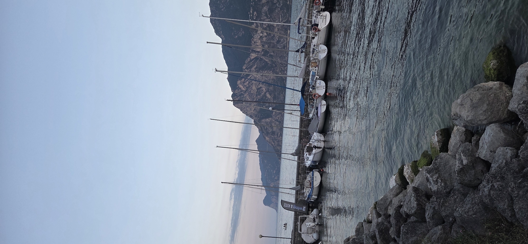 The image shows a marina in Torbole, Italy, with several sailboats docked along a stone pier. The boats are white with various colored sails and masts. In the background, mountains rise steeply from the water's edge, covered in vegetation. The sky is a pale blue with hints of pink, suggesting either sunrise or sunset. The water is calm, reflecting the boats and the sky. In the foreground, large rocks are piled along the shore. A lamppost is visible on the pier, and a banner with the text "WING & KITE" hangs from a pole. One small boat with two people is visible in the distance. The scene is serene and peaceful.