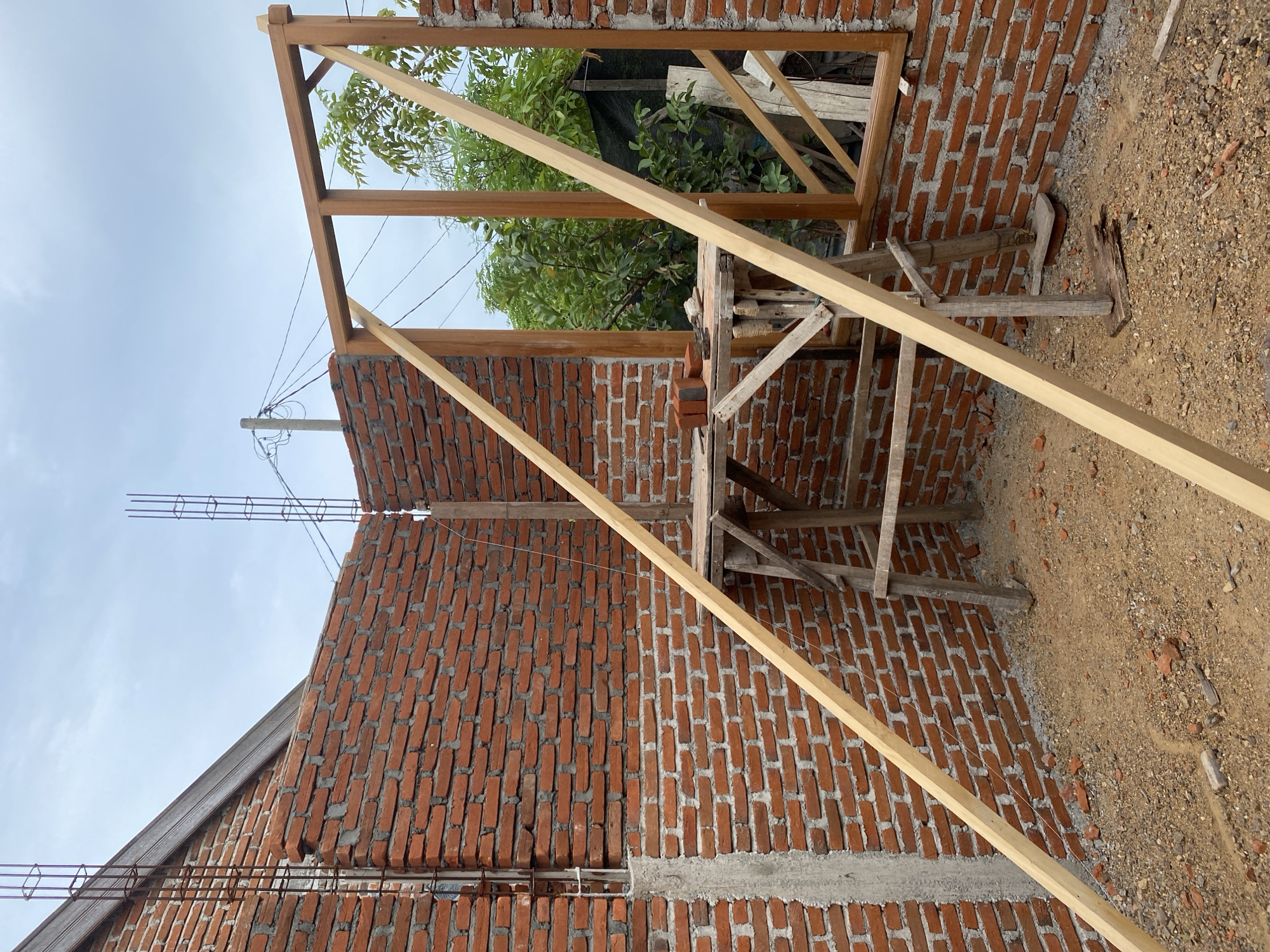 This image depicts an outdoor construction site in Banda Aceh, Indonesia, under a cloudy sky. The scene features a partially built brick wall, with exposed rebar protruding from a concrete pillar on the left. A wooden frame, presumably for a window or door, is erected within the brick opening. Two diagonal wooden planks are propped against the wall and the wooden frame, possibly serving as temporary support. A wooden scaffolding structure stands within the construction area, with a flat surface on top holding a few bricks and tools. The ground is covered with dirt, gravel, and scattered construction debris, including broken pieces of brick. In the background, lush green foliage is visible, suggesting the presence of trees and vegetation outside the immediate construction zone. Overhead, power lines and a utility pole are also present, indicating an urban or semi-urban setting. The overall impression is of an active construction project in progress.
