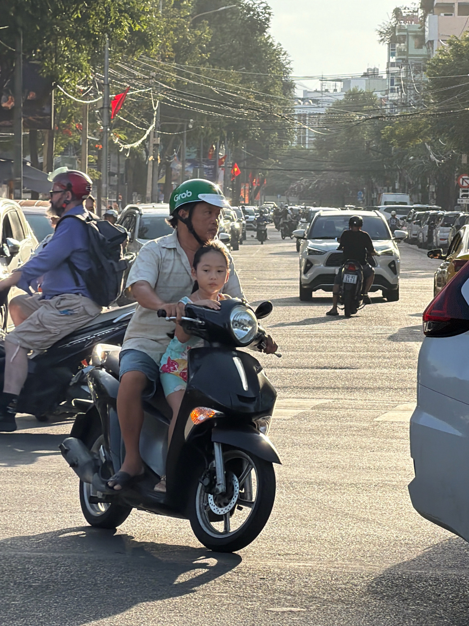 The image captures a street scene in Nha Trang, Vietnam, characterized by a bright, sunny day with a low angle of sunlight casting long shadows. The foreground features a dark scooter with a man and a young girl as passengers. The man, wearing a light-colored short-sleeved shirt and blue shorts, is operating the scooter. He has a green helmet with the "Grab" logo visible. The young girl, wearing a colorful top and light blue shorts, is seated in front of him. The scooter's front wheel, brake disc, and orange indicator light are visible.
To the left of the main scooter, another scooter is partially visible with a rider wearing a red helmet and a purple shirt, carrying a black backpack. The scooter is black.
The background is a busy street with multiple vehicles, including cars and numerous other scooters, indicating a bustling urban environment. Cars are lined up on the right side of the road, and more scooters are in motion in the distance. Buildings with multiple stories and trees line the street, with a dense network of overhead electrical wires creating a common urban visual element in Vietnamese cities. Red flags are visible on poles in the background, adding a touch of color to the scene. The road surface appears to be asphalt.