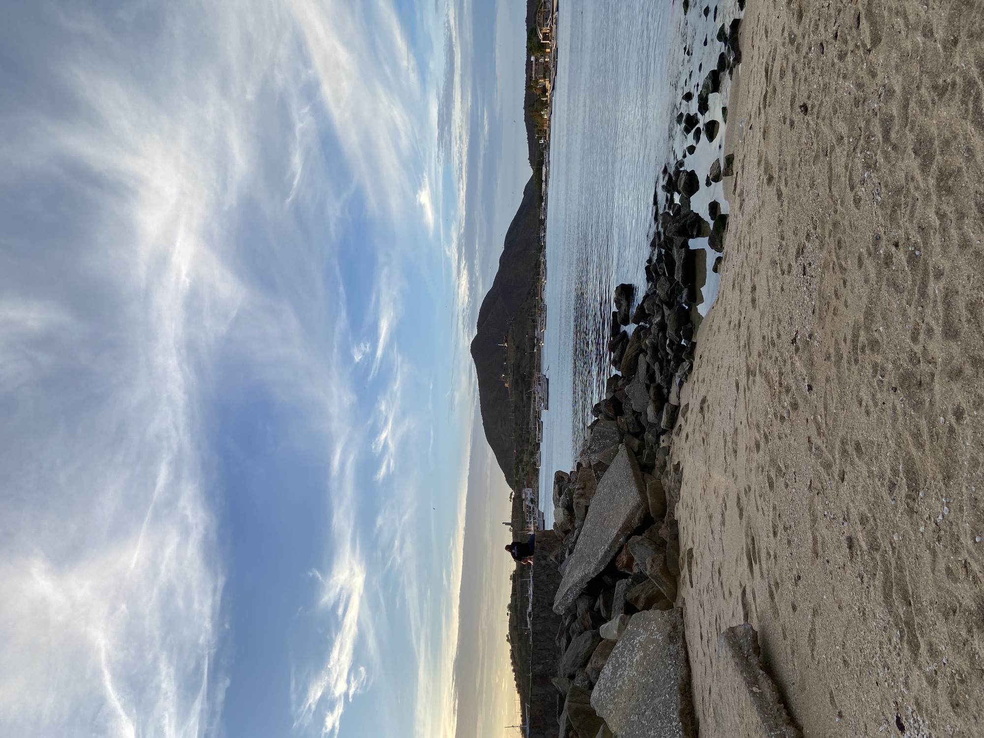 In Juan Griego, Venezuela, a serene coastal scene unfolds under a vast sky streaked with wispy white clouds against a gradient of soft blue. The foreground features a sandy beach littered with scattered seashells and the imprinted tracks of footprints. Adjacent to the beach, a rugged breakwater is formed by a jumble of large concrete slabs and dark rocks, some submerged in the calm, reflective water. A solitary figure is seated on the stone wall of the breakwater, their back to the viewer, gazing out at the bay. The bay itself is dotted with numerous moored sailboats and larger vessels, their reflections shimmering on the water's surface. In the distance, a chain of dark, hilly landforms stretches across the horizon, with scattered buildings and lights visible along the coastline, suggesting the presence of the town. The overall atmosphere is one of quiet contemplation and natural beauty.