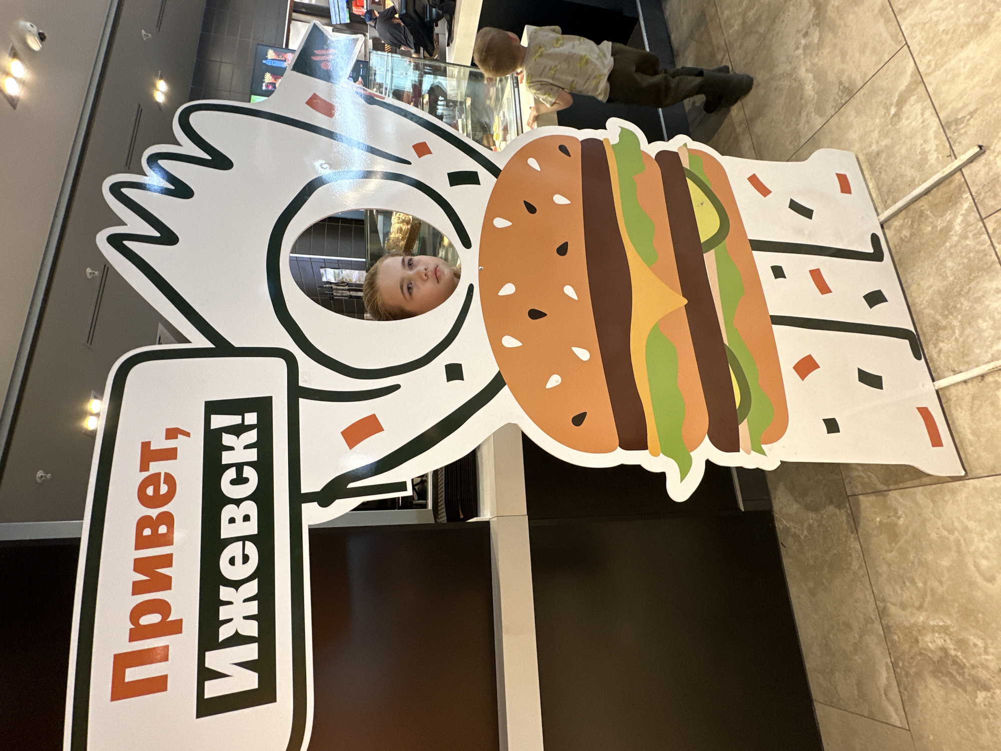 A child's face appears through a hole in a cartoonish burger cut-out. The cut-out has a sign that reads "Привет, Ижевск!" (Hello, Izhevsk!) indicating it is in the city of Izhevsk, Russia. Another child looks over the counter in the background. The floor is tiled.