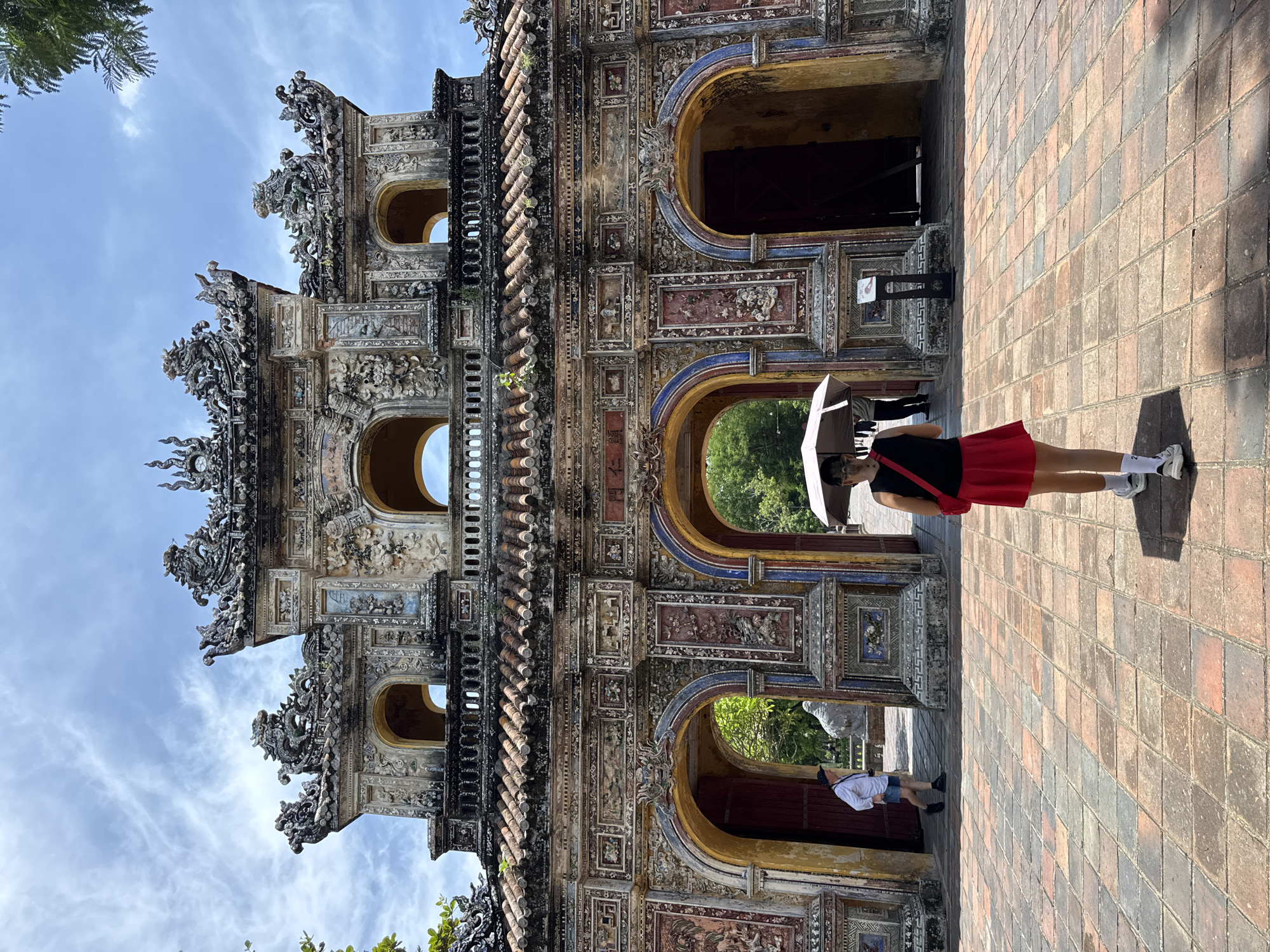 In Phú Xuân Ward, Vietnam, the Imperial City's Ngọ Môn (Meridian Gate) stands as a grand, ornate entryway. The gate, constructed with intricate details and adorned with dragon motifs, features multiple arches. In the foreground, a person walks towards the gate, holding a light-colored umbrella to shield themselves from the sun. They are wearing a black top, a red skirt, white socks, and sneakers. Behind them, another individual in a white shirt and blue shorts walks away from the gate. The ground is paved with square bricks in varying shades of red and orange. The sky above is a clear blue with scattered white clouds.