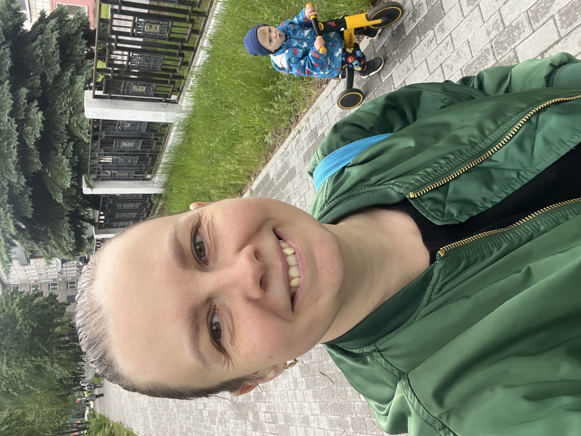 This media file is a rotated outdoor selfie featuring an adult woman and a young child in what appears to be a park or pedestrian area in Chaikovsky, Russia.
The woman, positioned in the foreground, is smiling broadly at the camera. She has light skin, brown eyes, and her hair is pulled back. She is wearing a dark green bomber jacket with a prominent gold zipper over a black top, with a light blue layer visible underneath. A small gold hoop earring can be seen in her right ear.
In the background, a young child is seated on a yellow and black balance bike or tricycle. The child is dressed in a blue patterned jacket, a blue beanie, dark pants, and black and white sneakers. The word "Bimboo" is visible on the bike's frame. The child is looking towards the camera, seemingly engaged with the person taking the selfie.
The scene includes a paved pathway constructed from light grey rectangular bricks, running alongside a strip of green grass. Beyond the grass, there is a dark metal fence with intricate, decorative panels. Tall evergreen trees are visible behind the fence, and in the far background, several multi-story residential buildings can be distinguished. The lighting is soft and even, suggesting an overcast daytime, indicative of mild weather. The woman's smile conveys a cheerful and pleasant emotion, capturing a happy moment between her and the child.