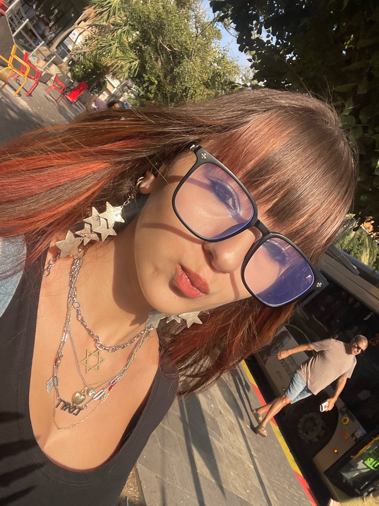 The image shows a close-up, high-angle selfie of a young woman with reddish-brown hair and bangs, wearing glasses and multiple layered necklaces, including one with a Star of David pendant. She is outdoors, with dappled sunlight suggesting a sunny day. In the background, trees and a light pole are visible. To the right and further back, a man wearing a light brown t-shirt and blue shorts is walking. He appears to be near a bus. The scene suggests an urban or suburban environment. The presence of the man and the bus implies this could be Raanana, Israel, based on common public transportation and demographics.