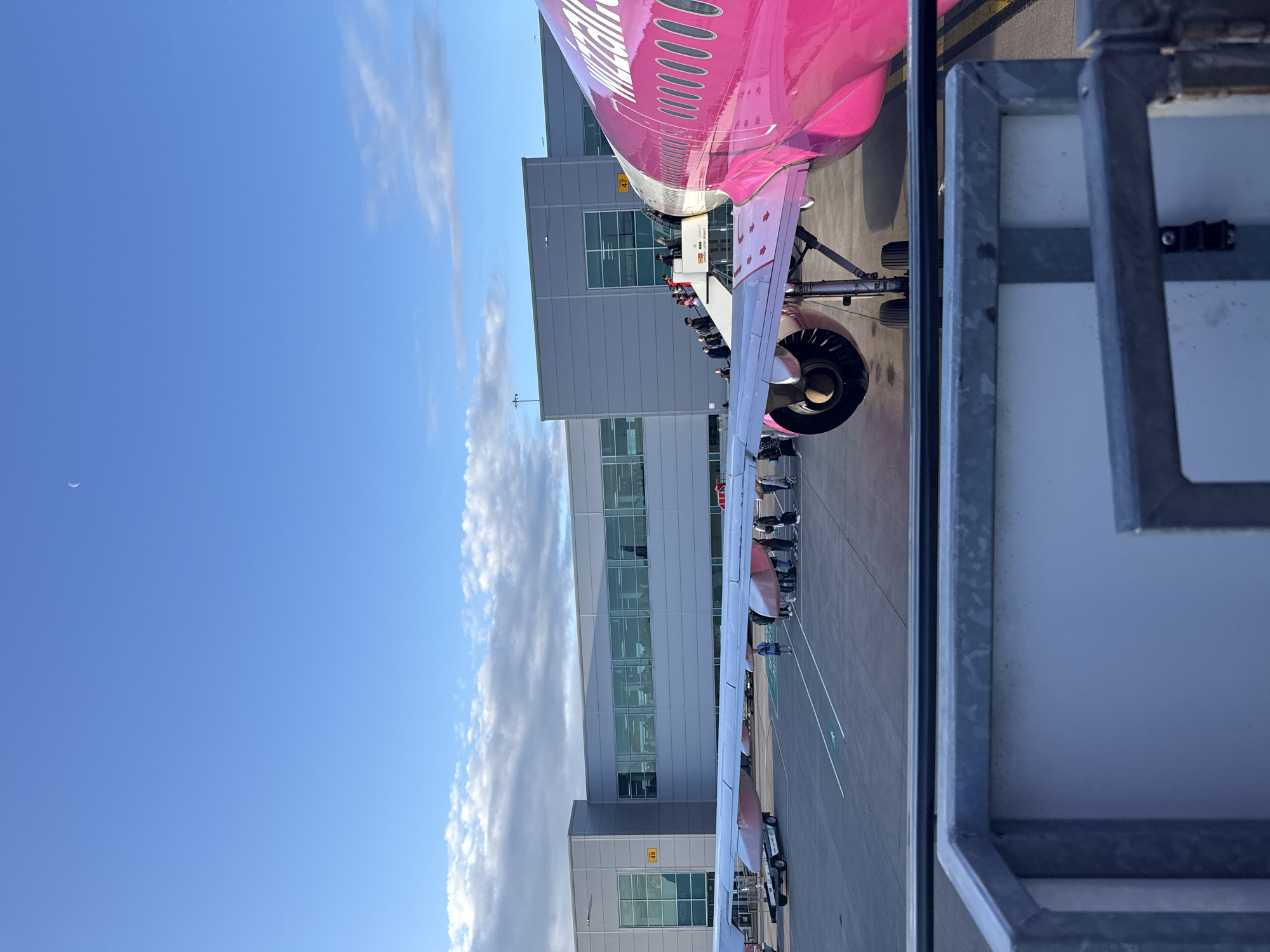 A pink airplane is parked at an airport gate. People are boarding the plane using stairs. The airport terminal is visible in the background. The sky is blue with some clouds. There is a crescent moon in the sky.