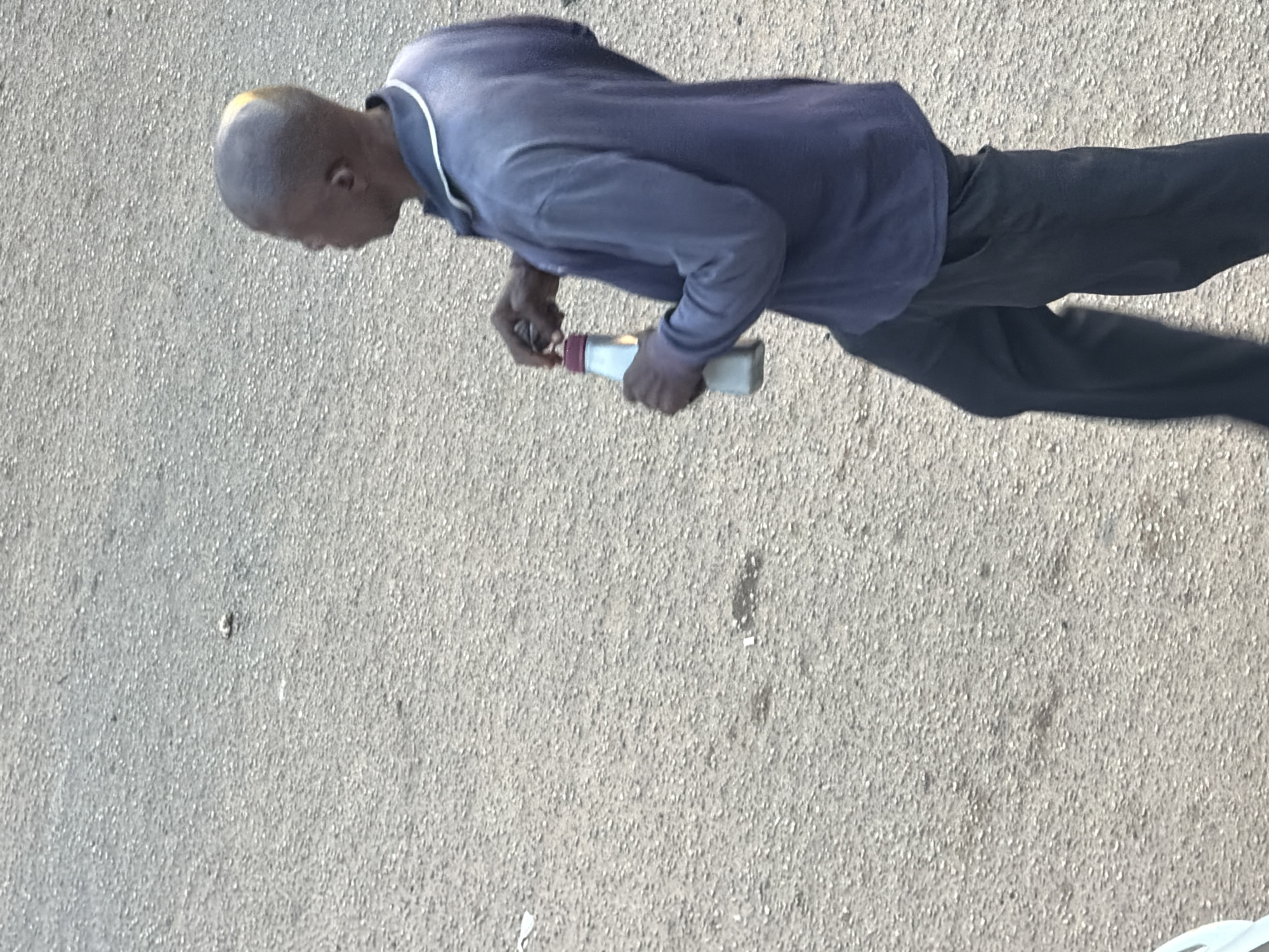 In Bukuru, Nigeria, a man with a shaved head and a yellow streak on the back of his head walks on an asphalt surface. He is wearing a blue long-sleeved collared shirt and black pants. He is holding a white and red bottle in his right hand and appears to be opening it. The ground is a light gray asphalt with visible pebbles.