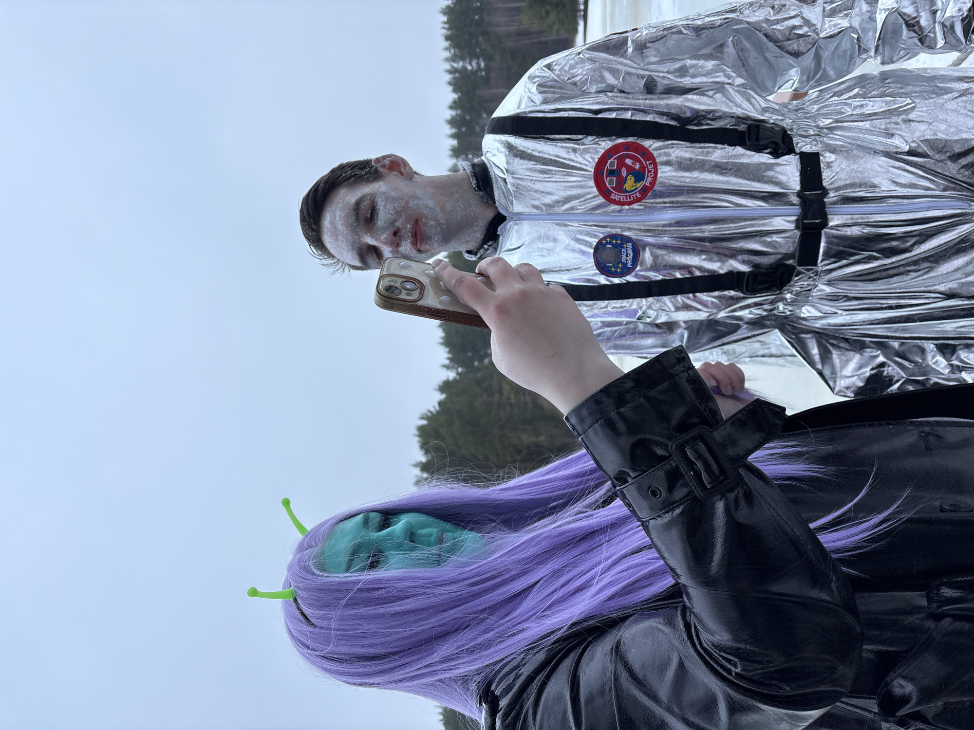 The image shows two individuals in costume near a snowy, possibly iced-over area, potentially the Kama River in Prikamskii, Russia. One person is dressed in a silver astronaut suit, with patches visible on the chest. His face appears to have white makeup on it. The other person, wearing a black leather jacket and a long purple wig with green antennae, has a blue-green painted face suggesting an alien character. The alien is holding a smartphone, possibly taking a photo of the astronaut. The background includes some bare trees and a cloudy sky.
