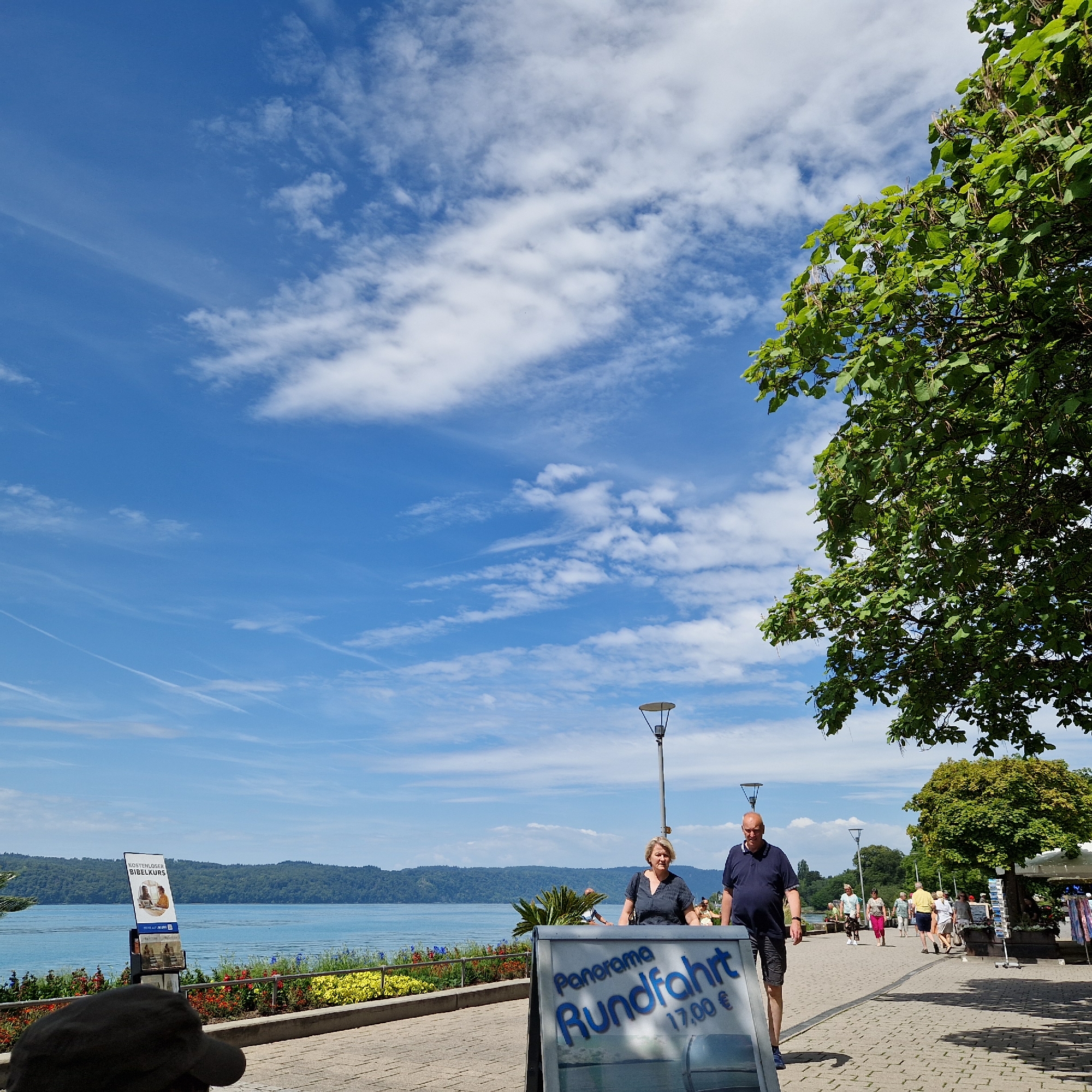 In Überlingen, Germany, a sunny day is captured along the waterfront promenade. The sky is a vibrant blue, dotted with scattered white clouds, with a large, leafy green tree dominating the right side of the frame. In the foreground, a sign advertises "Panorama Rundfahrt" for 17.00€. A man and woman stand near the sign, facing towards the camera and the lake beyond. The lake, a tranquil blue, stretches across the background, bordered by a lush green hillside. Several other people are visible walking along the promenade in the mid-ground, some near a small kiosk. The promenade itself is paved with interlocking stones. A smaller sign to the left advertises "KOSTENLOSER BIBELKURS" (Free Bible Course).