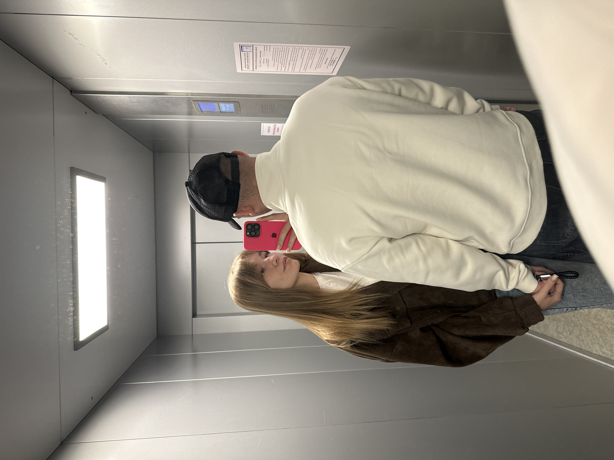 The image captures two individuals, a man and a woman, taking a mirror selfie inside a sleek, modern elevator with reflective metallic grey walls and ceiling. The perspective is slightly upward, showing the elevator's interior.
The man, primarily seen from the back, wears a light cream or off-white sweatshirt and a black mesh-back baseball cap. He holds a vibrant pink smartphone, its screen reflecting the woman's face and upper body. His left arm is bent, bringing the phone up for the photo.
The woman is visible in the phone's reflection. She has long, straight light brown hair with bangs, wearing a white top and a dark brown, possibly suede, jacket. She looks towards the camera with a calm expression.
Their hands are clasped together, with a black leather keyring visible in their joined grasp, suggesting an intimate connection.
Above the man's head, on the elevator wall, a digital display panel indicates "2" and an upward arrow, signifying the elevator is on the second floor and ascending. Next to the display, a vertical white paper notice with black text is affixed to the wall. While the specific text is unreadable, its format suggests it contains rules or instructions related to elevator use. Bright rectangular light panels illuminate the interior from the left wall and ceiling. The floor is also a matching metallic grey. The setting is indoors, brightly lit by artificial light, depicting a casual moment between the two individuals in Chaikovsky, Russia.