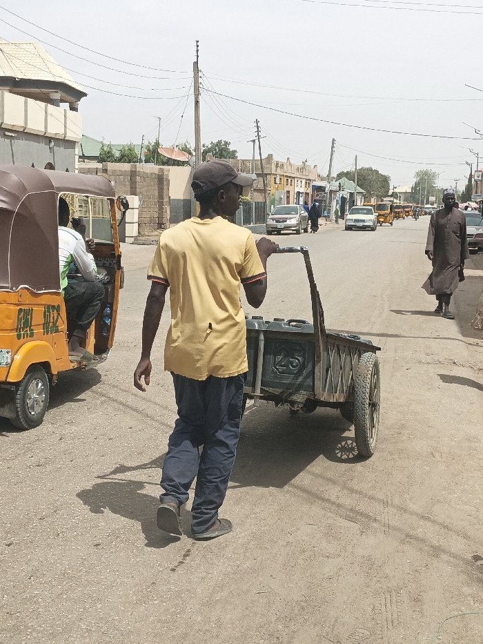 The scene depicts a daylight street view in Hotoro South, Nigeria. In the immediate foreground, a male individual, seen from behind, wears a brown baseball cap, a yellow short-sleeved t-shirt with a small vertical tear on the lower back, and dark blue sweatpants with white stripes on the side seams. He is pushing a dark-colored, two-wheeled wooden cart that contains at least two dark jerrycans, one visibly marked with "25". To his left, an orange three-wheeled auto-rickshaw (Keke Napep) with a dark brown canopy is parked or moving slowly. A passenger in a white and green shirt is visible inside, and a blue water bottle rests on the floor. A partial identification mark "GWL 10Z2" is present on the side of the Keke.
The setting is a paved road, transitioning to dirt or gravel shoulders, with visible tire tracks. Shadows on the ground indicate a high sun angle. Further down the street, on the right side, a male figure dressed in a long, brown traditional garment walks towards the background. Multiple vehicles, including a silver sedan, a white sedan, and several other auto-rickshaws, are present along the street. Several pedestrians are also visible in the mid-distance.
Buildings, primarily one and two stories, line both sides of the road, constructed from concrete or brick, exhibiting various states of finish and color. A prominent building on the left features a light-colored, multi-pitched roof. Utility poles with numerous overhead power lines are visible throughout the background. A large red satellite dish is mounted on a rooftop in the mid-left. The sky is a hazy light blue.
