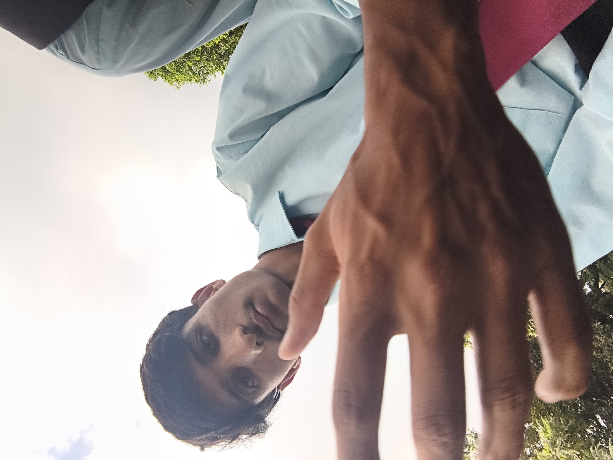 A low-angle, close-up, outdoor selfie depicts a young man with dark hair and a mustache, looking towards the camera. He is wearing a light blue collared shirt and a pink tie. His right hand is held up in the foreground, partially obscuring his face. The background shows a bright sky with some clouds, green foliage, and possibly trees. The image appears to be taken in a casual setting, possibly in Dharamshala, India, given the natural elements and attire visible.
