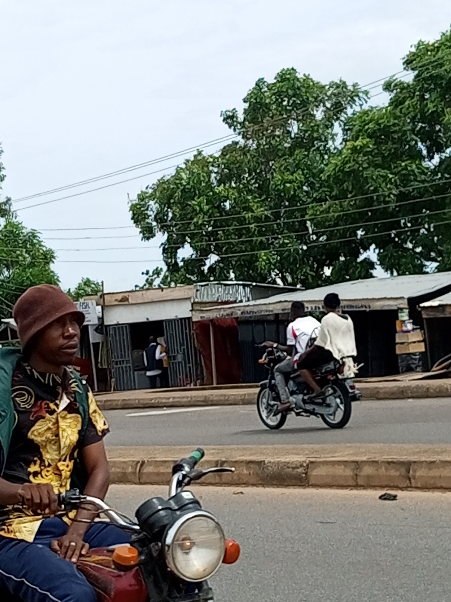 The image, taken on a street in Garko, Nigeria, captures a street scene with multiple motorcycles and roadside shops. In the foreground, a person wearing a brown beanie and a patterned shirt is riding a motorcycle. Their hands are on the handlebars, and they are looking to the left of the frame. Behind them, another motorcycle is visible with two passengers. The passenger in front is wearing a white shirt with red accents, and the passenger behind is wearing a white garment. They are riding away from the camera. The background features a row of modest shops with metal grills on their windows and entrances. Trees with dense green foliage line the upper part of the image, and power lines are visible against the overcast sky. The road surface is asphalt, with a concrete curb separating it from the area where the shops are located. The overall impression is of a busy, everyday street in a Nigerian town.