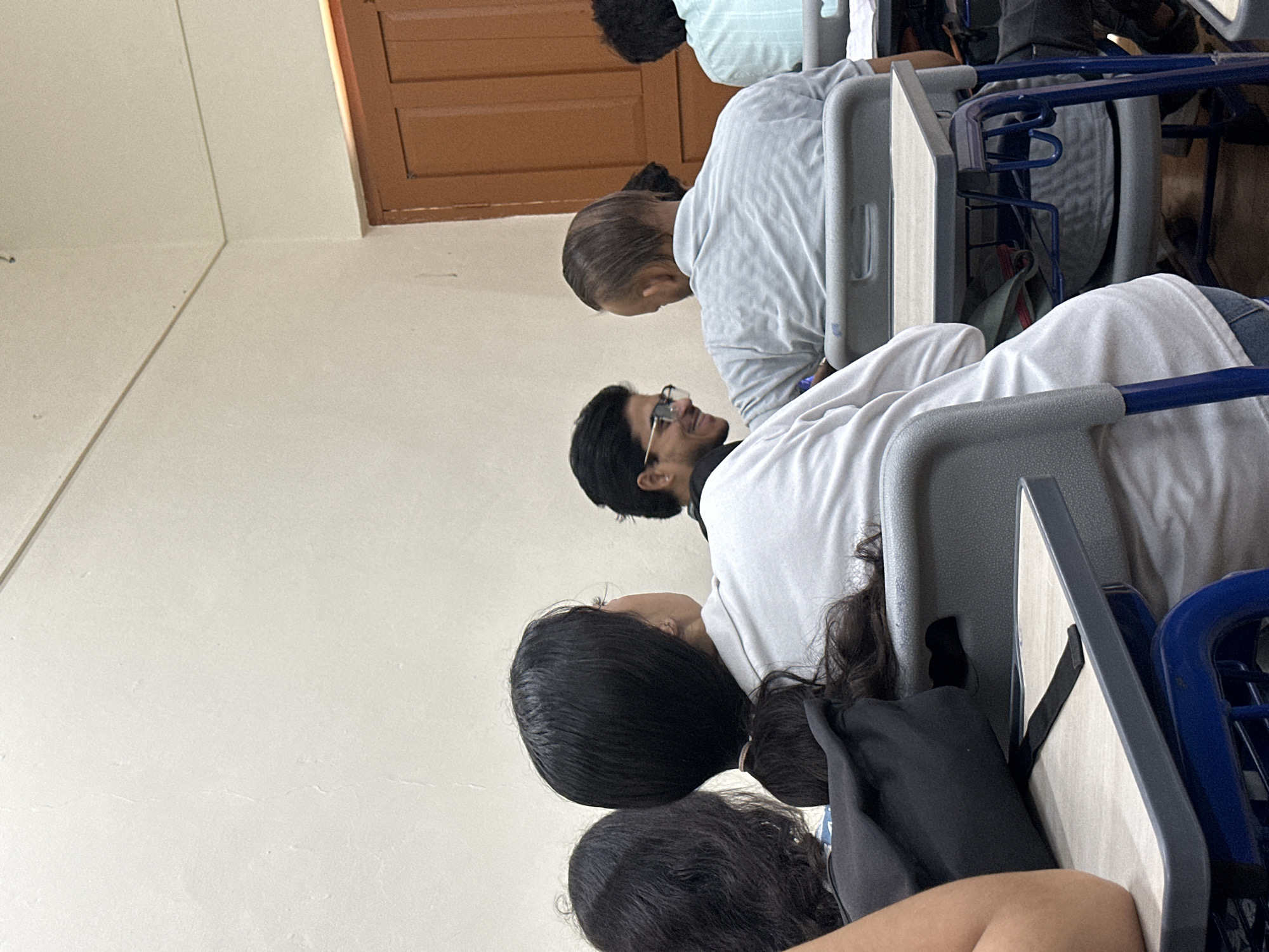 A medium shot captures an indoor scene with several individuals seated at desks, likely in a classroom or lecture hall. The perspective is from behind and slightly to the side of a person with dark hair pulled into a ponytail, wearing a light-colored top. To their left, another person with dark, wavy hair is visible from the side. In front of the ponytail-haired individual, a person with dark hair and glasses is looking to their right, smiling. To the right of the ponytail-haired individual, a person with receding dark hair and a light-colored shirt is also seated. Further to the right, another individual in a light-colored shirt is partially visible. The environment appears to be a room with off-white walls and a wooden door in the background. Desks with blue frames and light-colored tabletops are arranged in rows. A black bag is placed on the floor next to the ponytail-haired person's desk. The lighting suggests an indoor setting, possibly during the day.