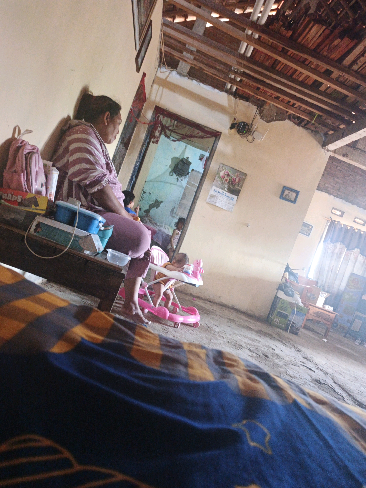 The image depicts an indoor scene, likely a home, in Bumiharjo, Indonesia. In the foreground, a woman in a striped pink shirt and purple pants is seated on a wooden bench. She is looking to her right. Beside her on the bench are a pink backpack, a yellow box, and blue containers. To her right, a baby is in a pink baby walker. In the background, a doorway leads to another room, and a calendar with flowers is hanging on the wall. The room has a concrete floor and a partially exposed wooden ceiling. The overall environment appears modest and functional.