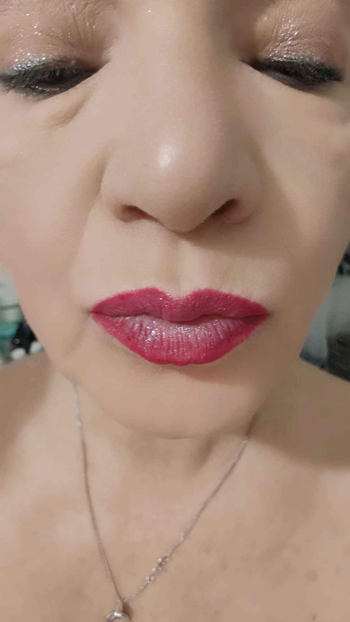 A close-up photograph focuses on the face of a person with their eyes closed. The person has dark, lined eyes and appears to be wearing shiny eye shadow. The nose is centrally positioned and dominates the image. The lips are painted a vibrant shade of red. A thin, silver necklace with a pendant is visible around the person's neck. The environment appears to be indoors and the background is out of focus. The city of Krasnoyarsk, Russia, is not discernible from the details in the image.