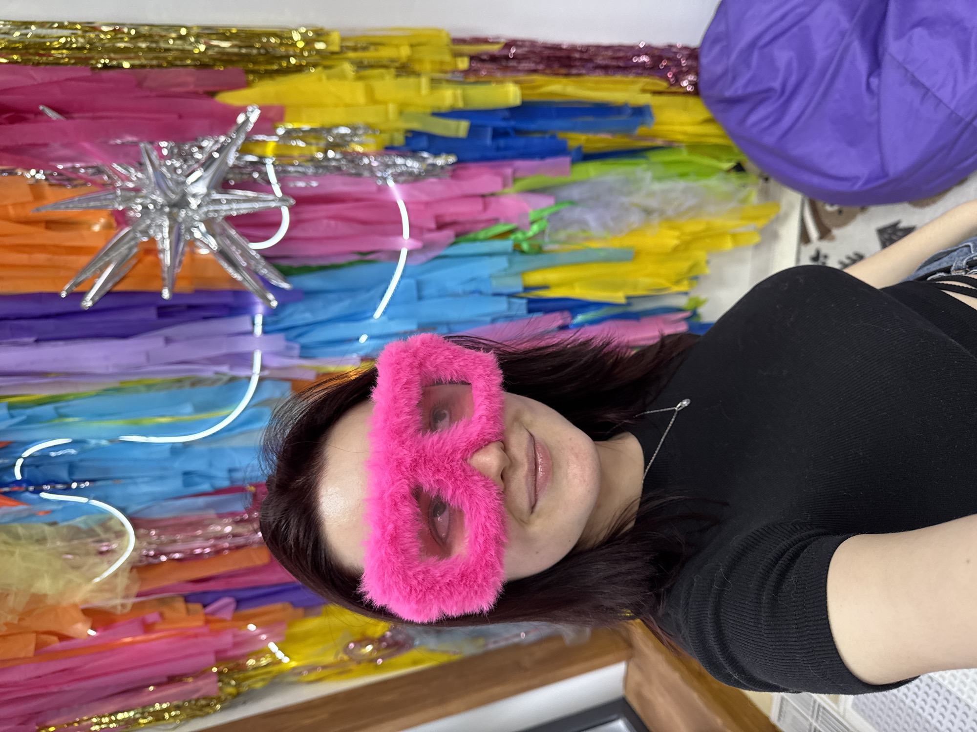 A person wearing a pink fuzzy eye mask poses in front of a colorful backdrop. The backdrop consists of hanging streamers in various colors, including gold, silver, yellow, pink, orange, blue, green, and purple. A silver star-shaped ornament is attached to the backdrop. A purple beanbag chair is visible in the upper right corner of the image. The person is wearing a black shirt and a silver necklace.
The city of Izhevsk, Russia, cannot be determined from the image.
