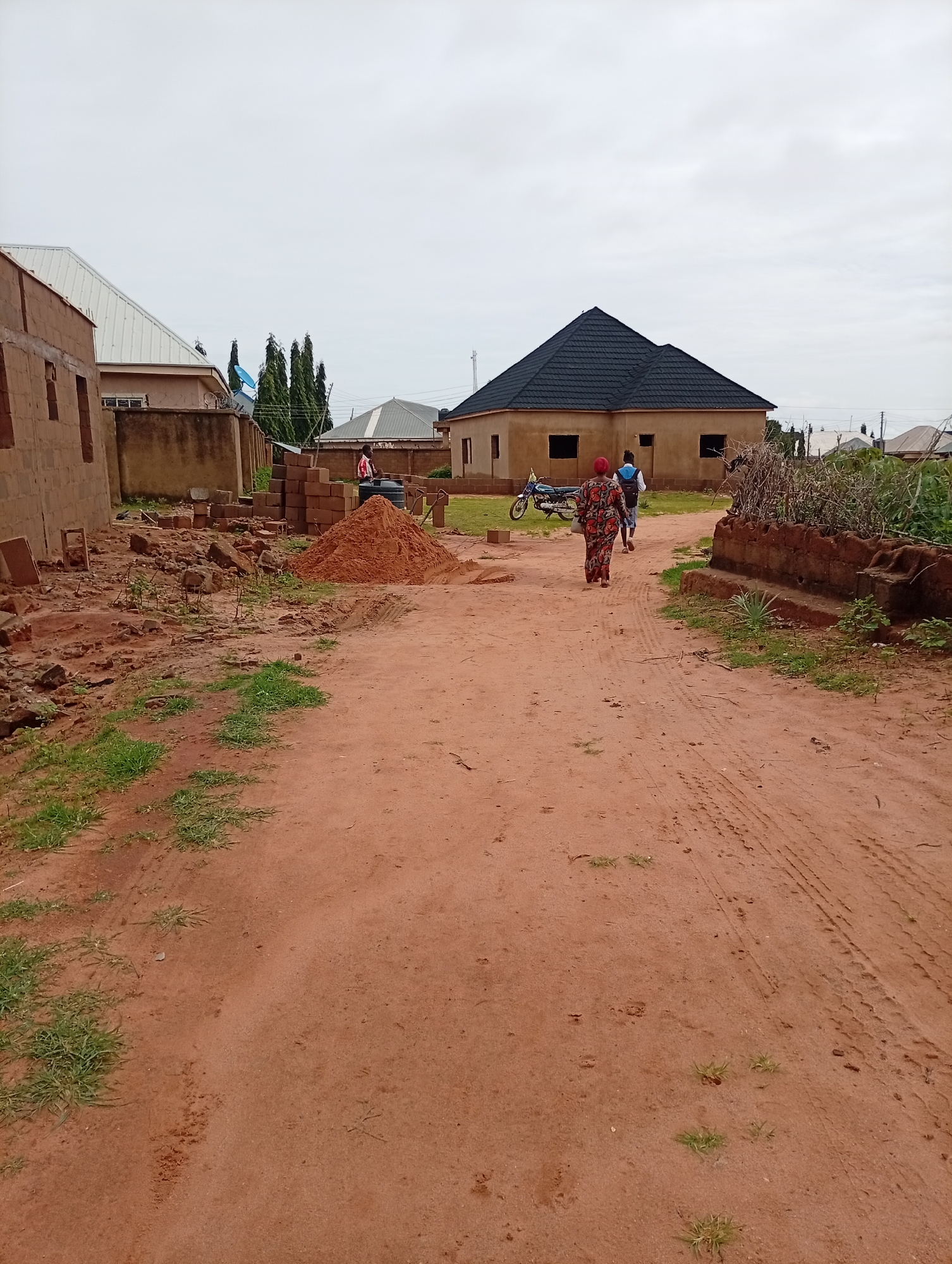 The image depicts an outdoor scene on a dirt road in Tumfure, Nigeria, under an overcast sky. In the foreground, a reddish-brown dirt road is visible, with tire tracks and patches of sparse green grass growing along its edges.
In the middle ground, two individuals are walking away from the camera down the road. The person on the left wears a colorful, patterned dress and a headscarf. The person on the right wears a blue shirt and white shorts. To their left, a motorcycle is parked. Further left, construction materials are piled up, including bricks and a large mound of sand. Several unfinished buildings are also present, constructed from brick and concrete. One building in the middle ground has a dark, angular roof and visible window openings.
In the background, more buildings are visible, some with corrugated metal roofs. A few tall, slender trees stand behind the buildings, and a blue satellite dish is mounted on a structure. The overall impression is a developing residential area with ongoing construction.