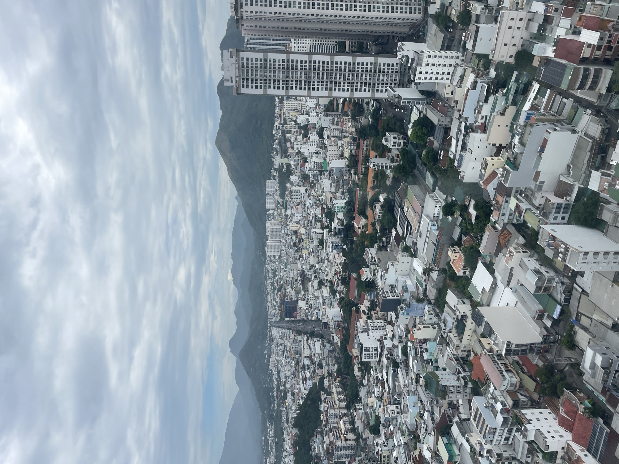 This is an aerial view of a dense urban landscape with mountains in the background. The city is characterized by a mix of low-rise buildings with red and grey roofs, as well as several modern high-rise apartment buildings. A wide, tree-lined road runs through the center of the city, leading towards the mountains. The sky is overcast, with a soft, diffused light illuminating the scene. The overall impression is one of a bustling city nestled within a natural landscape. This scene is consistent with an elevated view of Nha Trang, Vietnam, a coastal city known for its beaches and surrounding mountains.