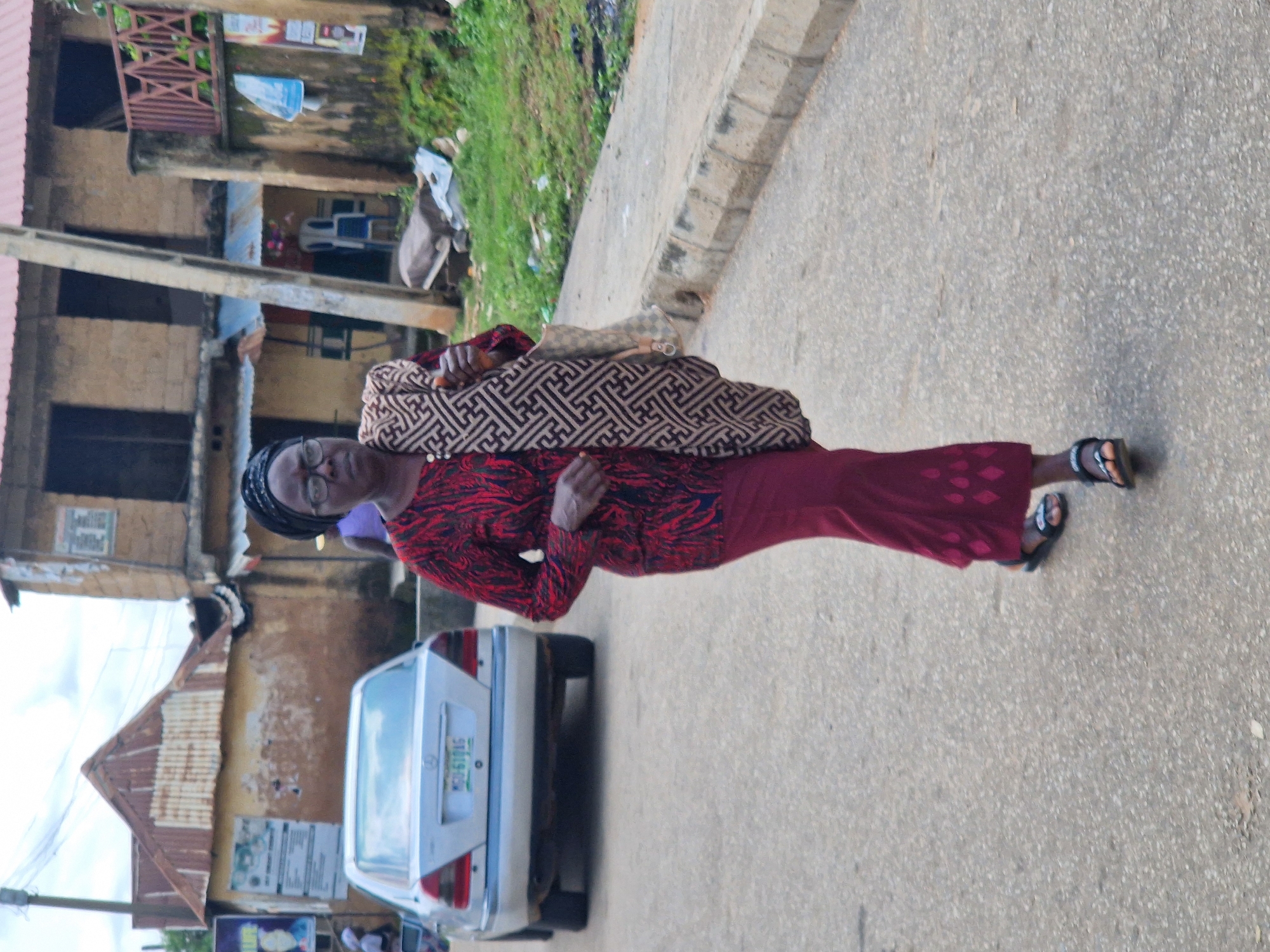 An outdoor, medium-shot, eye-level, full-body photograph shows a woman walking toward the viewer on a paved road in Kabong, Nigeria. The woman, who appears to be in her 60s, has dark skin, glasses, and is wearing a dark headscarf, a long-sleeved red and black patterned shirt, and a maroon skirt. She is carrying a light brown bag with a black geometric pattern slung over her left shoulder. Her sandals are black and her toenails are painted red. Behind her, a silver Mercedes-Benz sedan is parked on the left side of the road, facing away from the viewer. A two-story building with a red roof is visible in the background, with a lighter colored facade and dark windows. A utility pole stands to the right of the building. Green foliage and some debris are scattered along the right side of the road. The sky is overcast.