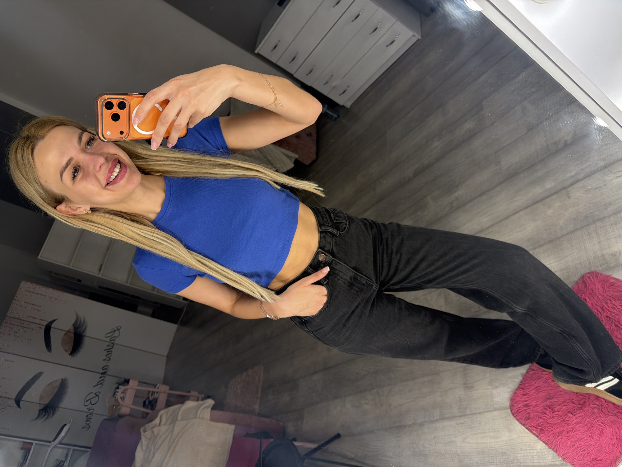 A young blonde woman is standing in what appears to be a room with a wooden floor and a large mirror. She is wearing a blue crop top and dark wash jeans. She is holding an orange smartphone in her right hand, taking a selfie. Her left hand is resting in her front pocket. In the reflection of the mirror, there is a pink rug on the floor, a rolling cart, and a piece of furniture with drawers. There is also a wall decor with stylized eyelashes. The overall impression is that of a private space, possibly a home or a beauty salon. There are no visible indicators that this scene is located in Kyiv, Ukraine.