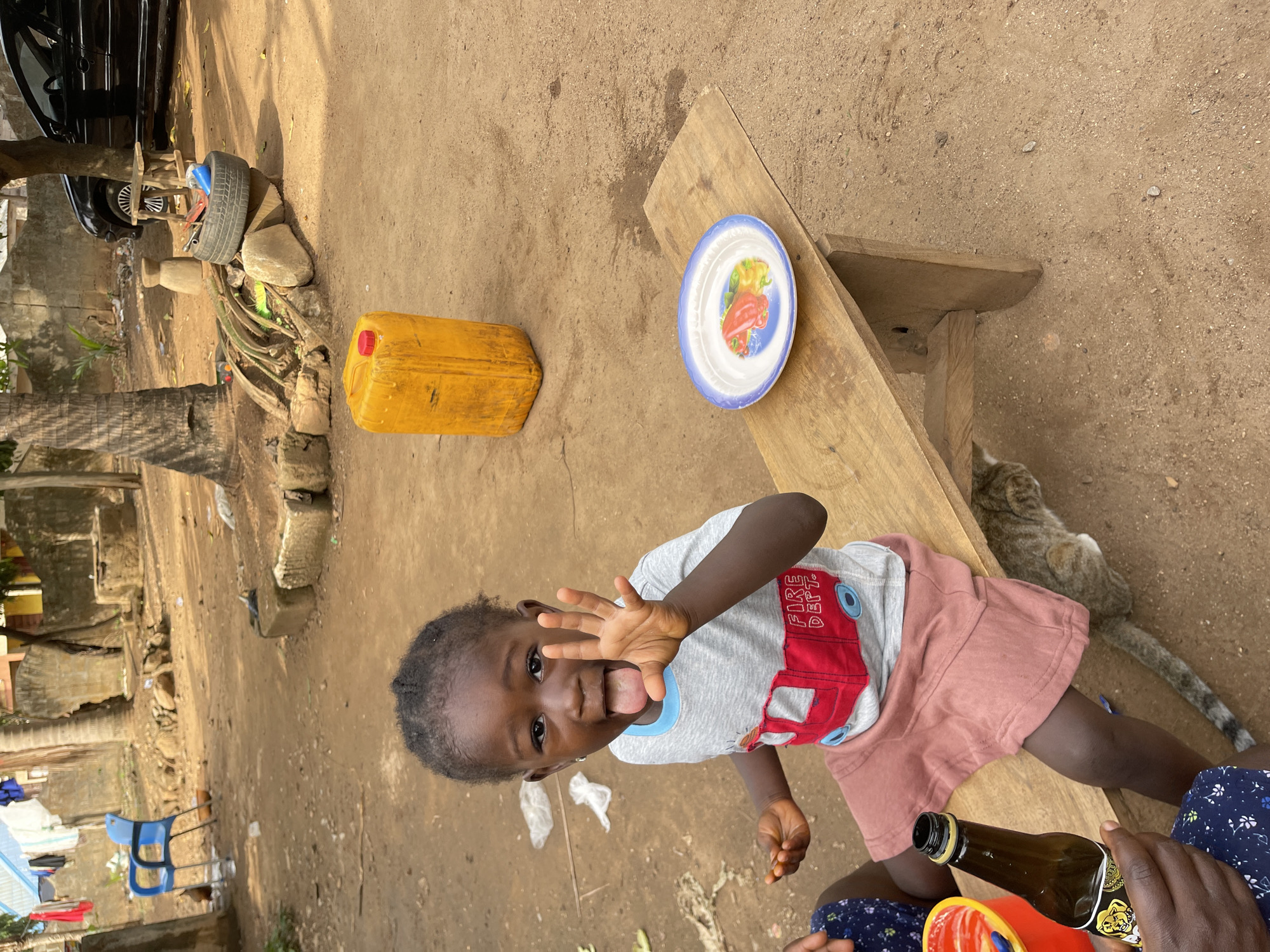 The image, likely taken in Akwanga, Nigeria, shows a child with dark skin, wearing a light gray shirt with a red fire truck design and pink shorts. The child is sticking their tongue out and holding up their hand, facing the camera directly. The background consists of a dirt surface, a wooden plank bench with a colorful plate on top, a yellow jerrycan, and a gray tabby cat resting on the ground near the bench. A brown bottle is visible in the lower right corner, held by a person wearing a blue floral patterned fabric. In the distance, there is a glimpse of residential buildings, laundry hanging on a line, and a blue plastic chair. A car tire and car are also visible in the top left part of the image.