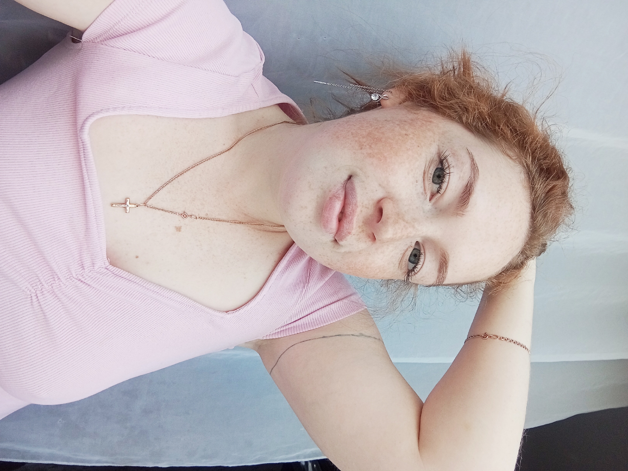 A fair-skinned young woman with red hair and freckles is lying down, presumably in Yurgamysh, Russia. She is wearing a pink shirt and a necklace with a cross pendant. A silver earring with a chain hangs from her left ear. She has a thin line tattoo on her arm and is wearing a bracelet on her left wrist. Her head is resting on a light blue or white surface.