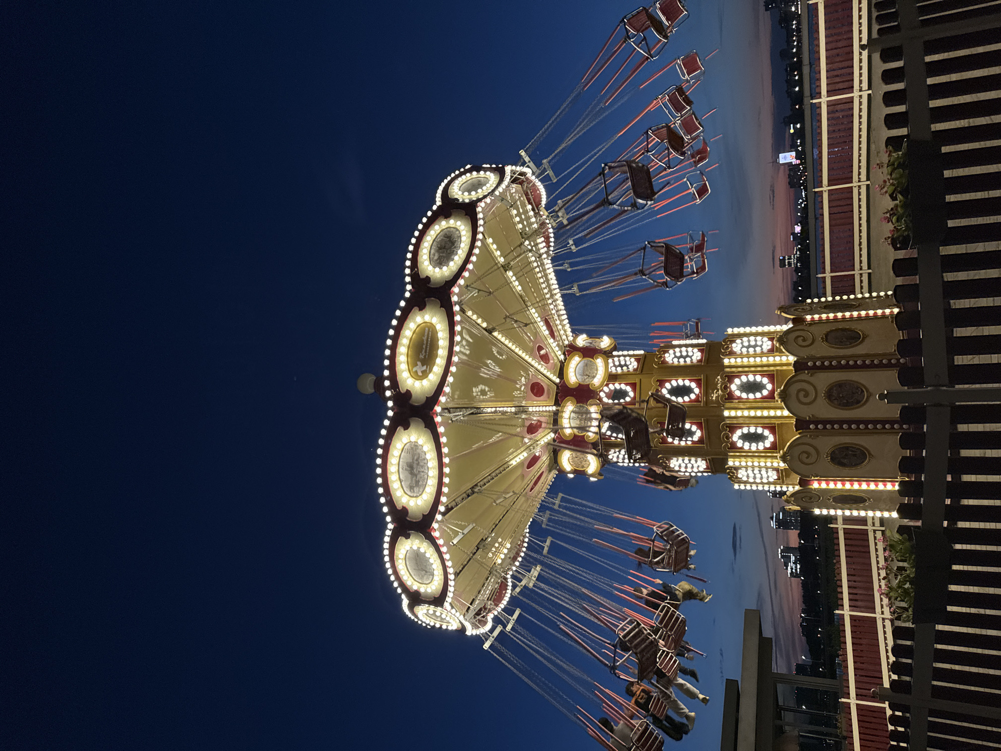 The image shows a fairground swing carousel in Kazan, Russia, taken at night. The carousel is brightly lit with many lights, with empty or occupied seats hanging from chains. The background features a deep blue evening sky with a faint horizon line of city lights. A wooden structure and some greenery are visible on the right side of the image, providing context to the carousel's location. The photo is taken from a low angle, emphasizing the height and grandeur of the carousel.