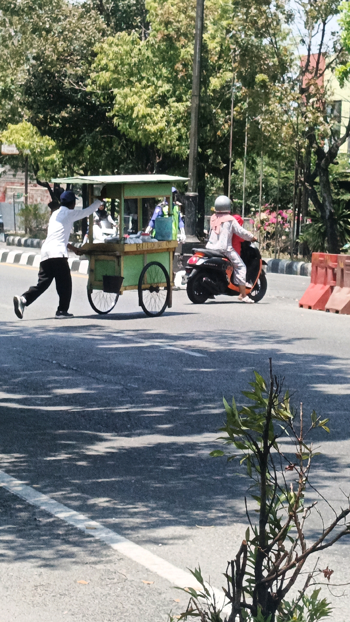 In the city of Surakarta, Indonesia, on a sunny day, a street vendor is seen pushing a light green food cart with two wheels along the side of a road. The cart has a light-colored canopy and appears to contain various items. A person wearing a white long-sleeved shirt, dark pants, and a dark cap is pushing the cart. In the background, a person is riding a scooter with an orange and black body, wearing a light-colored outfit and a silver helmet. The scooter is traveling in the same direction as the food cart. There are trees and buildings visible in the background, along with traffic barriers. The road surface is asphalt, with a white line marking a lane. Sunlight casts shadows on the road from the trees and foliage.