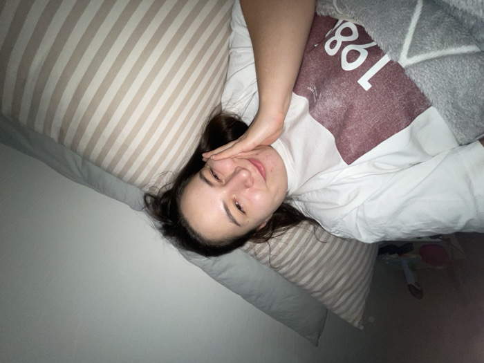 A person lies in bed with a striped pillow and blanket. They wear a white shirt with the number 1986 on it. Their hand is near their face as they smile into the camera. The room is dimly lit, showing a corner of the room with a glimpse of objects on the floor.
