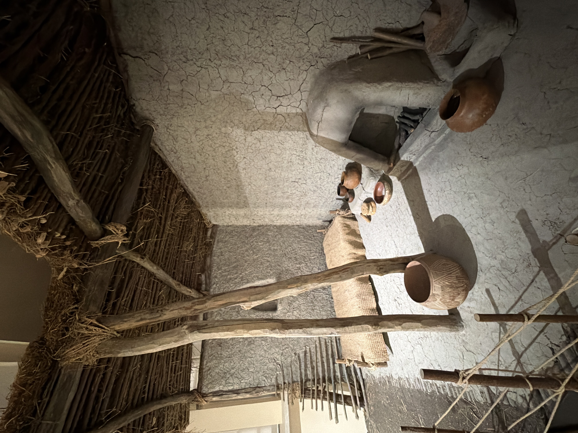 The image depicts a reconstructed interior scene, likely a diorama or exhibit, possibly located in a museum in Sofia, Bulgaria. The scene suggests an ancient dwelling or living space.
The walls appear to be constructed of a textured material, resembling dried mud or clay. A portion of the structure features a thatched or woven roof made of twigs and reeds. Several clay pots and vessels are scattered throughout the scene, along with a woven mat or textile.
There is a human figure, likely a static model, shown bending or working. Additional tools and implements are visible, further suggesting a domestic or occupational setting. There is also a rudimentary wooden ladder or structure. The overall environment is dimly lit, enhancing the aged and historical atmosphere.